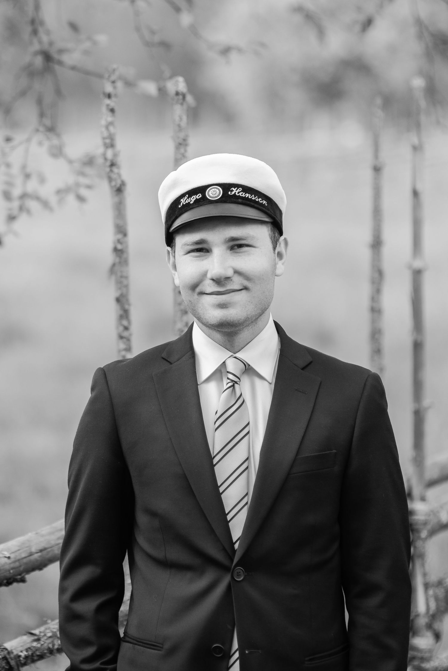 A person wearing a white cap and navy suit stands in front of a wooded background in black and white.