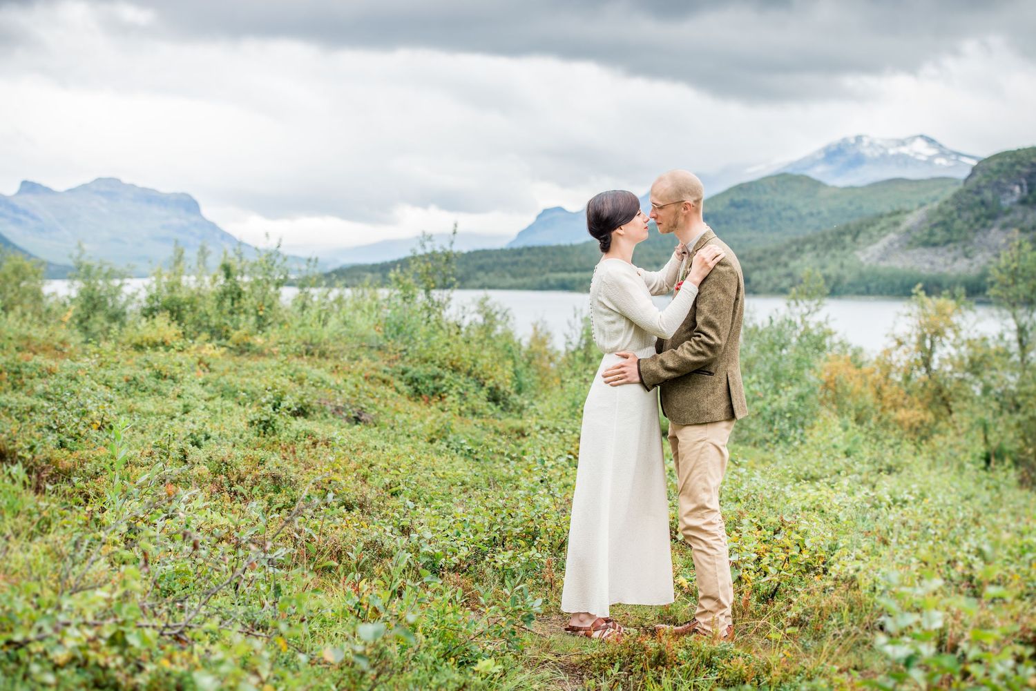 Two people embrace on a grassy hillside with mountains and lake in the background.