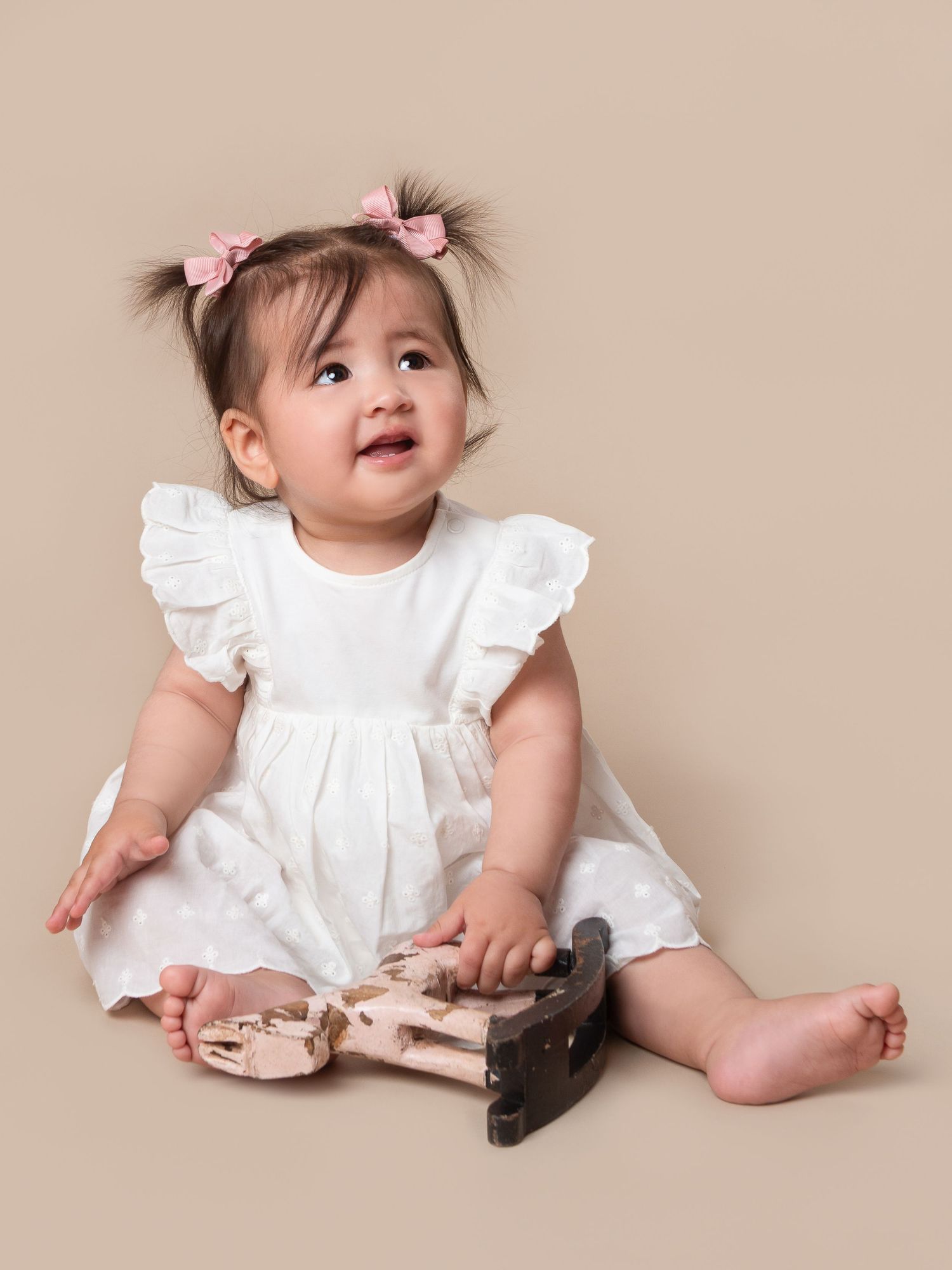 A cute baby girl in a white ruffled dress with pink hair bows sits on a beige background holding a wooden toy.