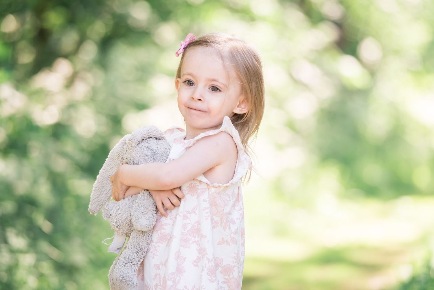 Young girl in pink floral dress hugging a stuffed bunny toy outdoors in a lush green garden setting.