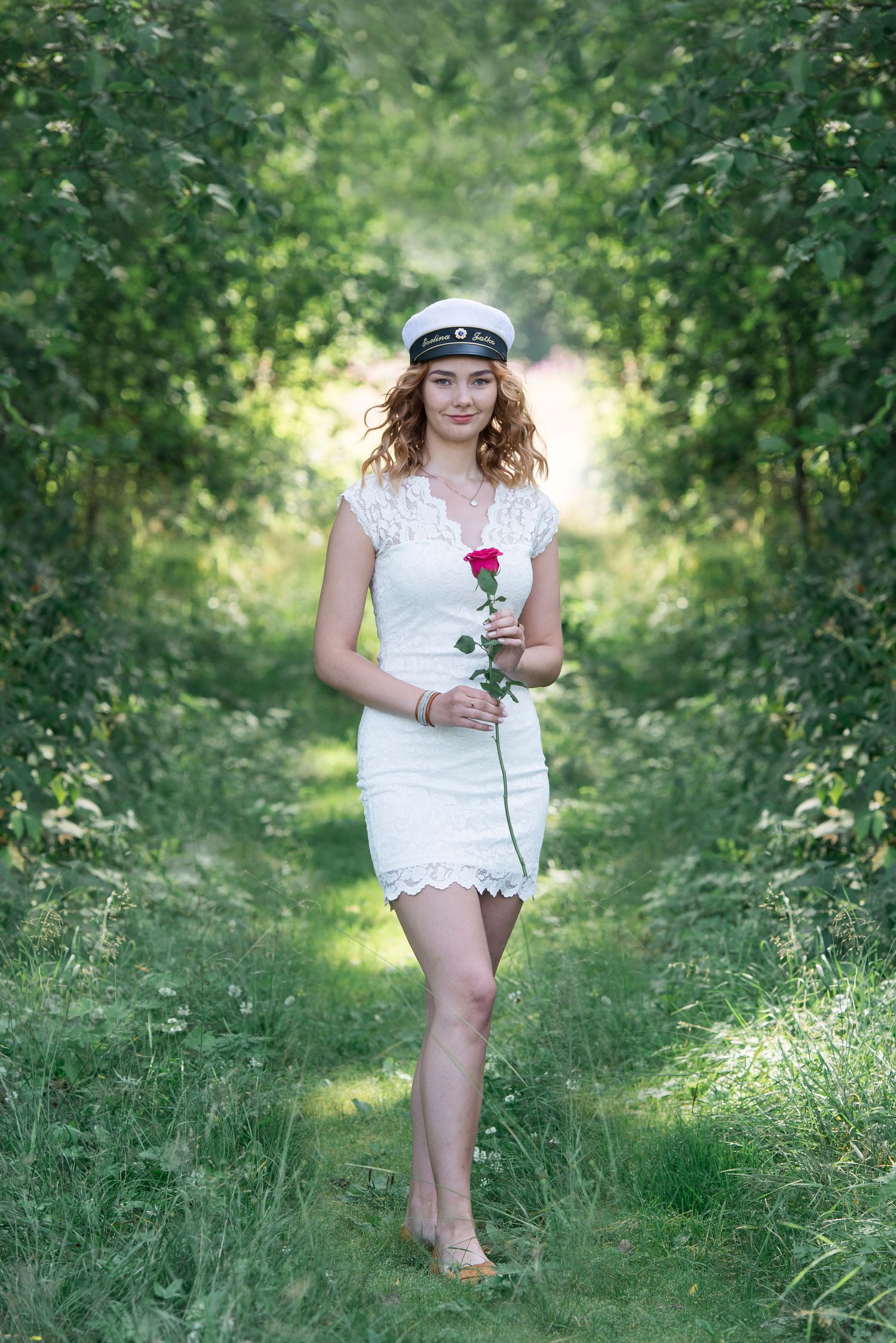 Someone in white lace outfit holding a red rose in a green garden path.