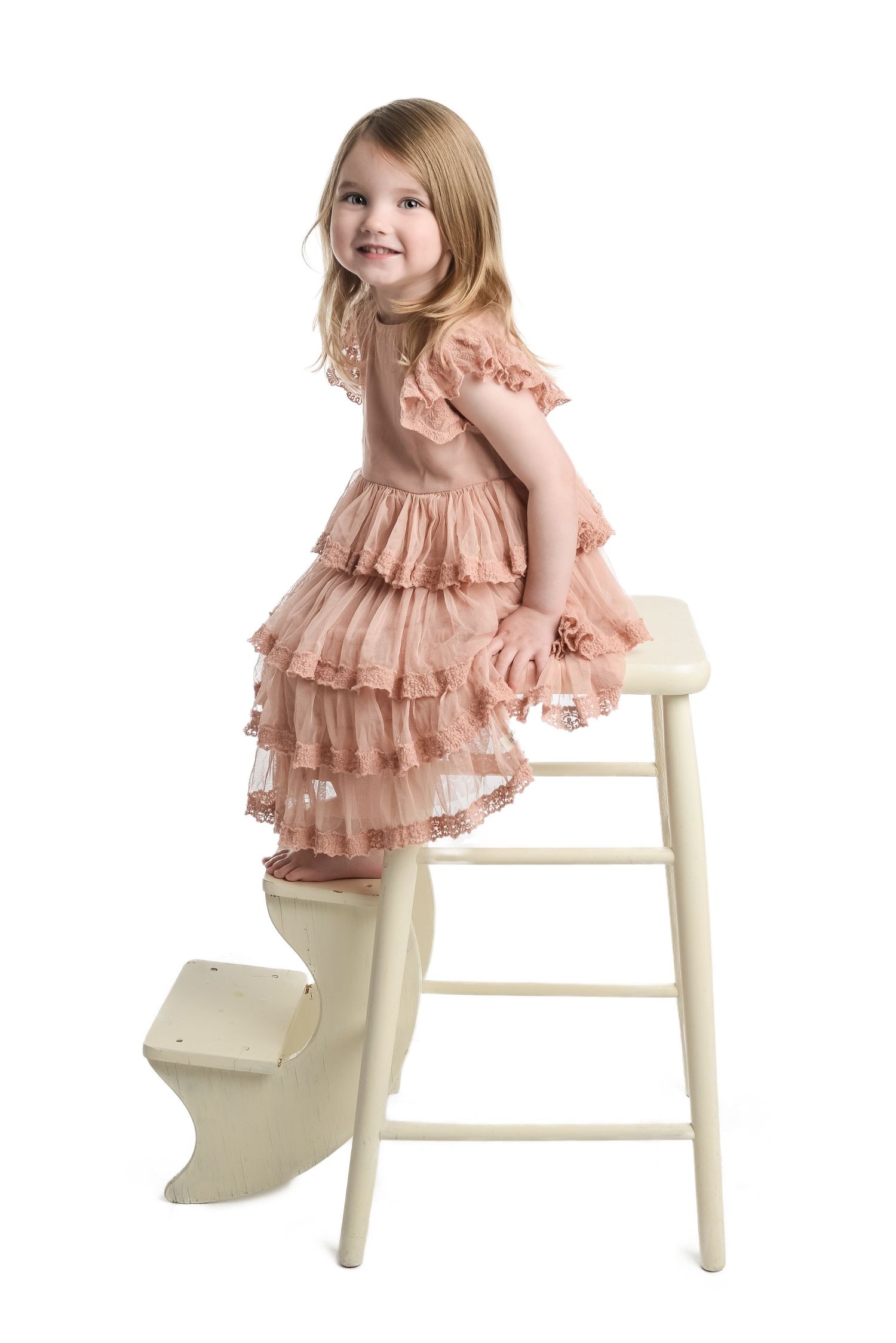 Young girl in pink layered dress posing on white wooden chair.