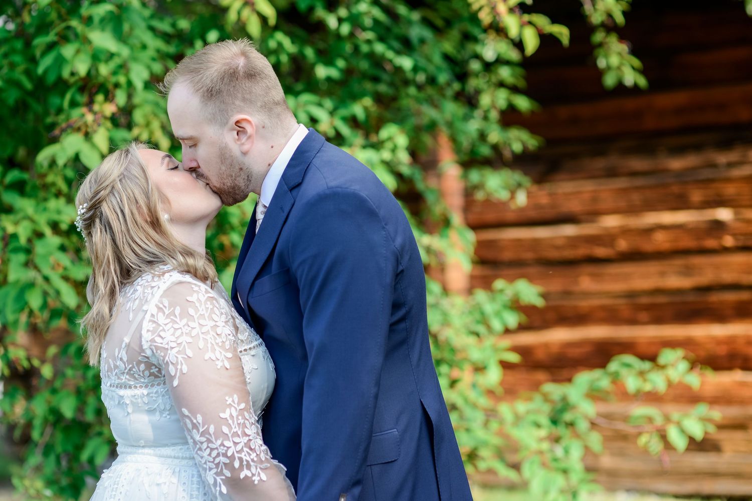 Wedding couple poses against rustic log cabin backdrop surrounded by green foliage.