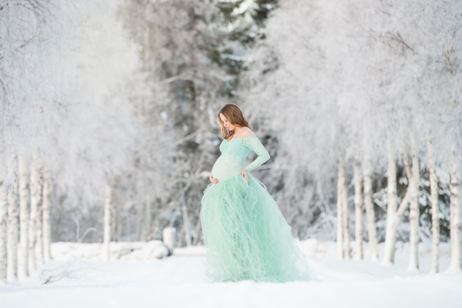 Pregnant woman in mint green tulle gown posing in snowy winter forest for maternity photo session.