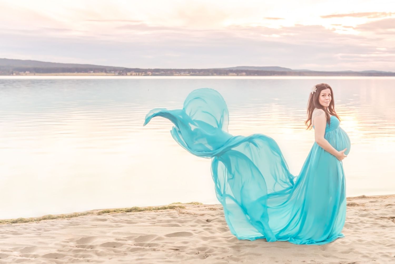 Pregnant woman in flowing teal gown on sandy beach at sunset with sheer fabric billowing in the wind.