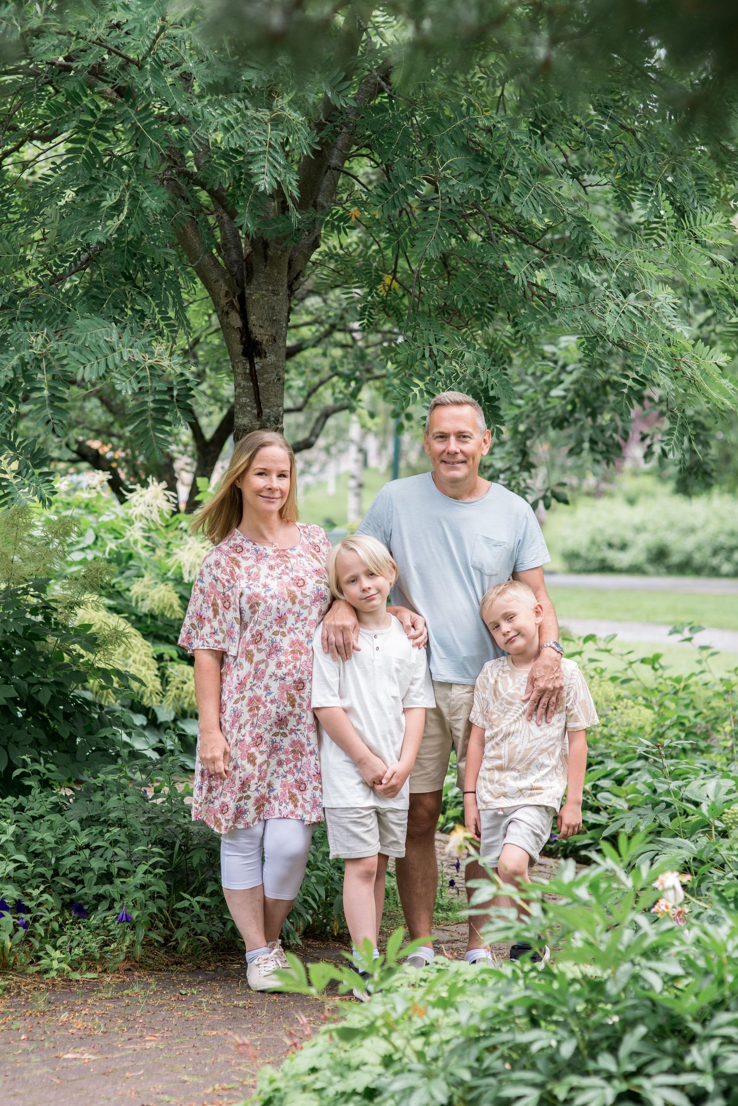 Family of four posing together on a garden path surrounded by lush green trees and plants on a summer day.