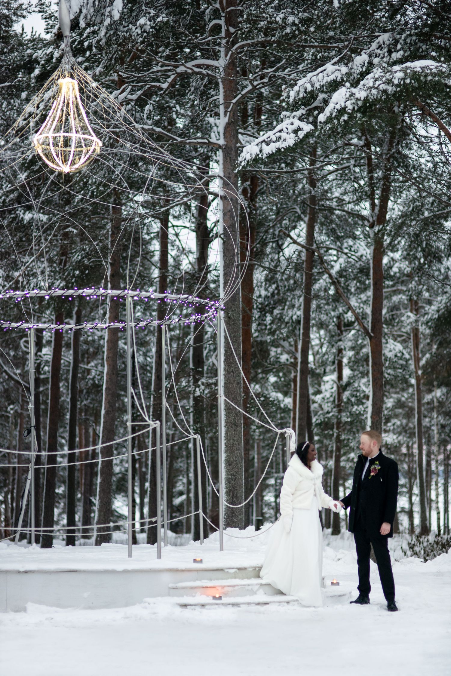 Winter wedding ceremony setup in a snowy forest with string lights and decorative elements.