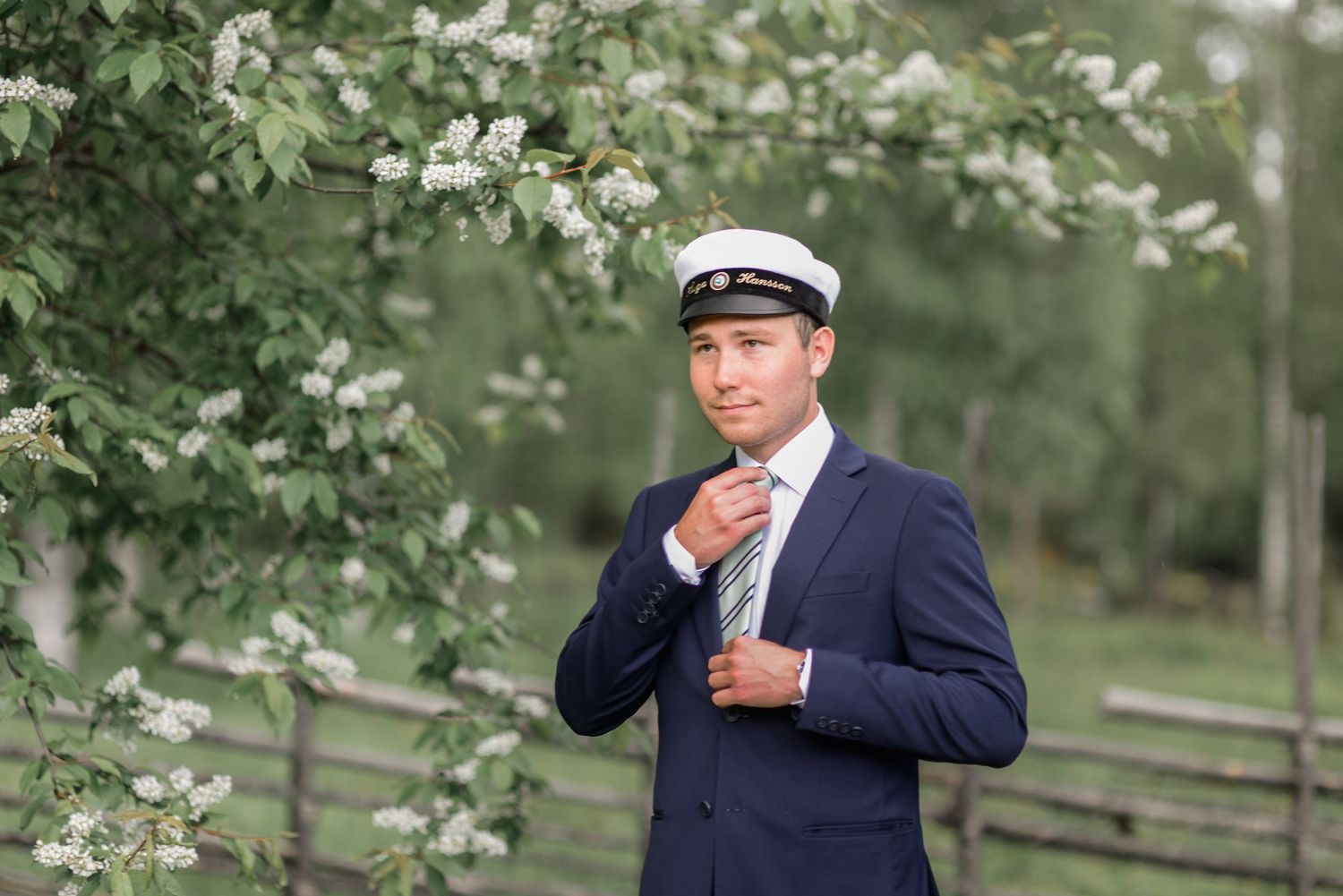 A person in a navy suit and captain's hat poses against a spring background with white blossoms.