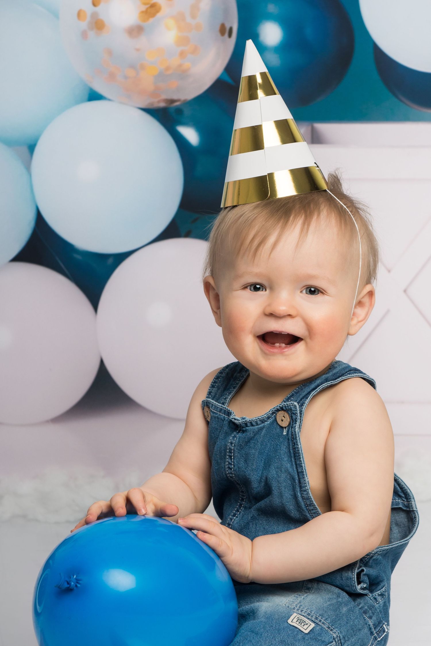 Blue balloon first birthday photoshoot with denim overalls and party hat decorations.