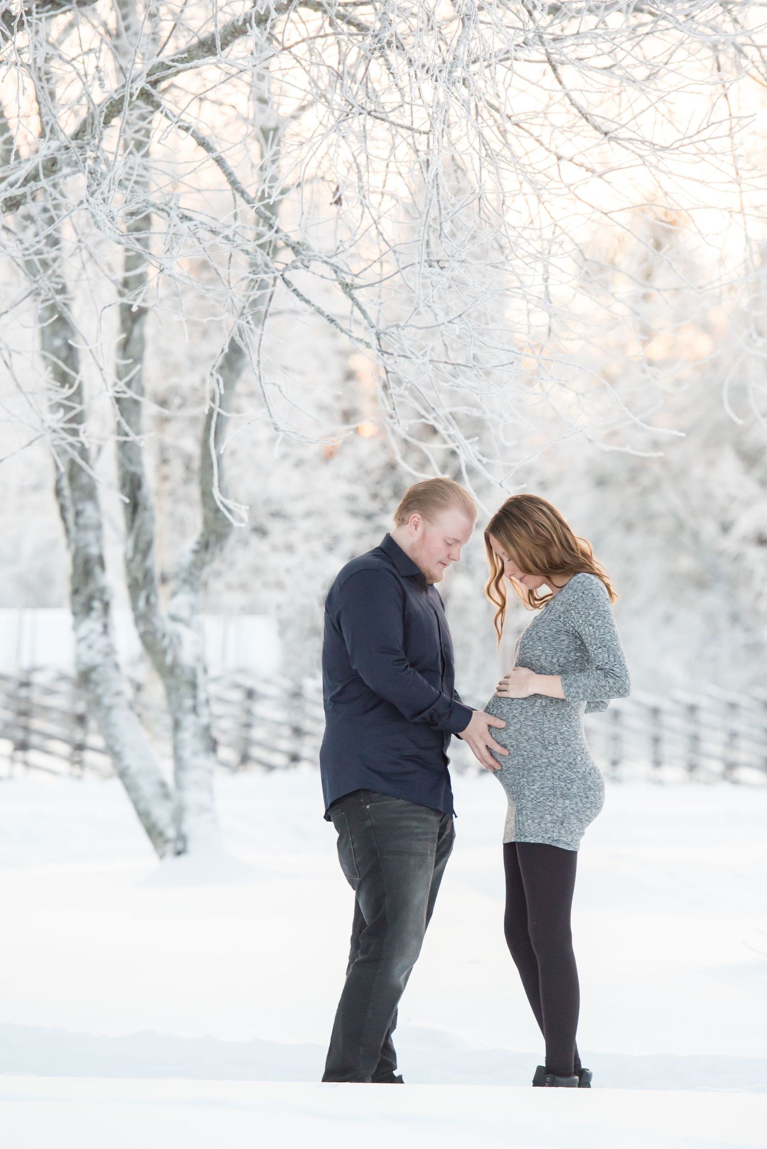 Couple sharing a tender moment in a snowy winter landscape, woman in gray dress showing baby bump.