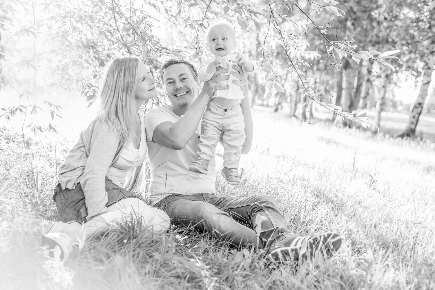 Black and white photo of happy family sitting in grass, parents lifting smiling baby outdoors in park setting.