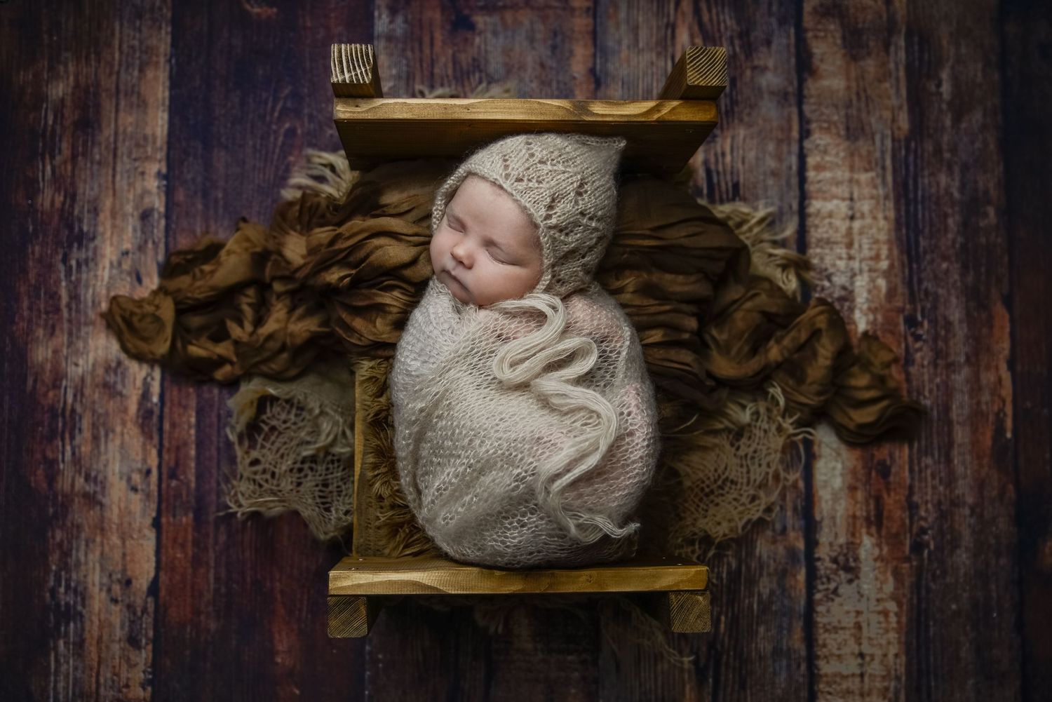 Newborn wrapped in beige swaddle sleeping in rustic wooden frame with textured brown backdrop.