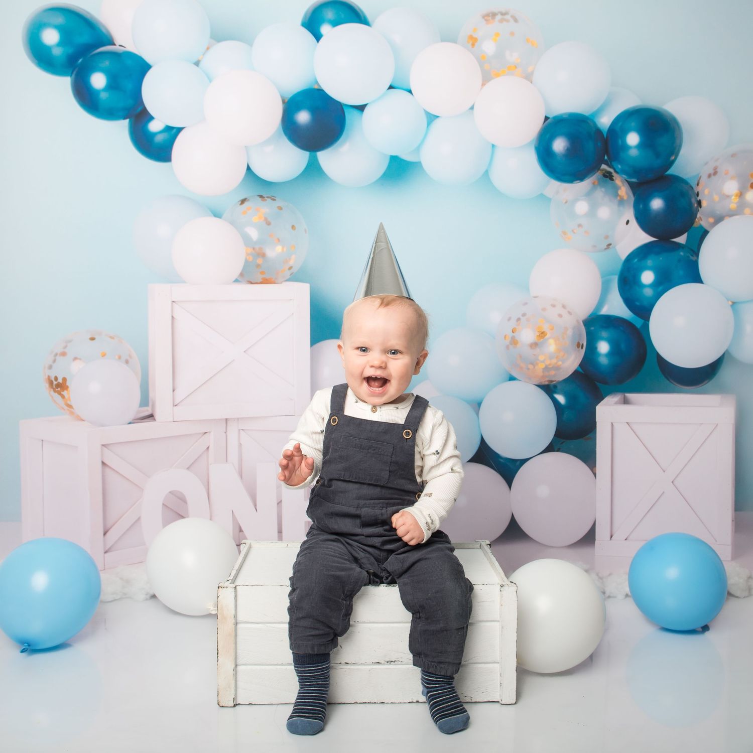 Festive first birthday setup with blue and white balloons against white backdrop for cake smash photoshoot.