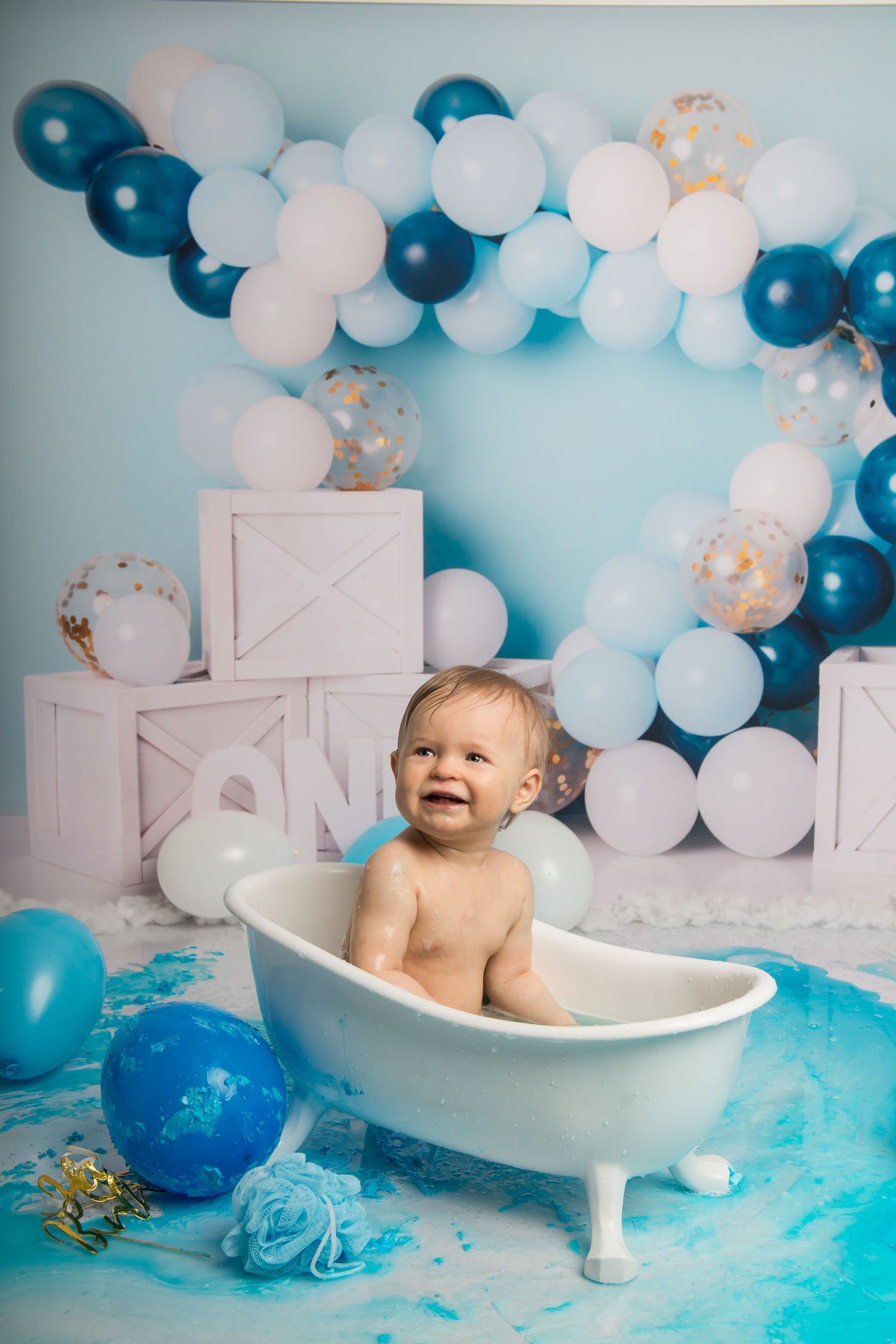 A vintage-style bathtub surrounded by blue and white balloons in a cake smash photography setup.