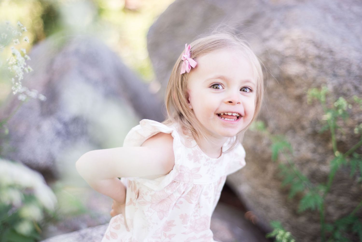 A joyful toddler girl in a floral dress with a pink bow smiles outdoors near large rocks.