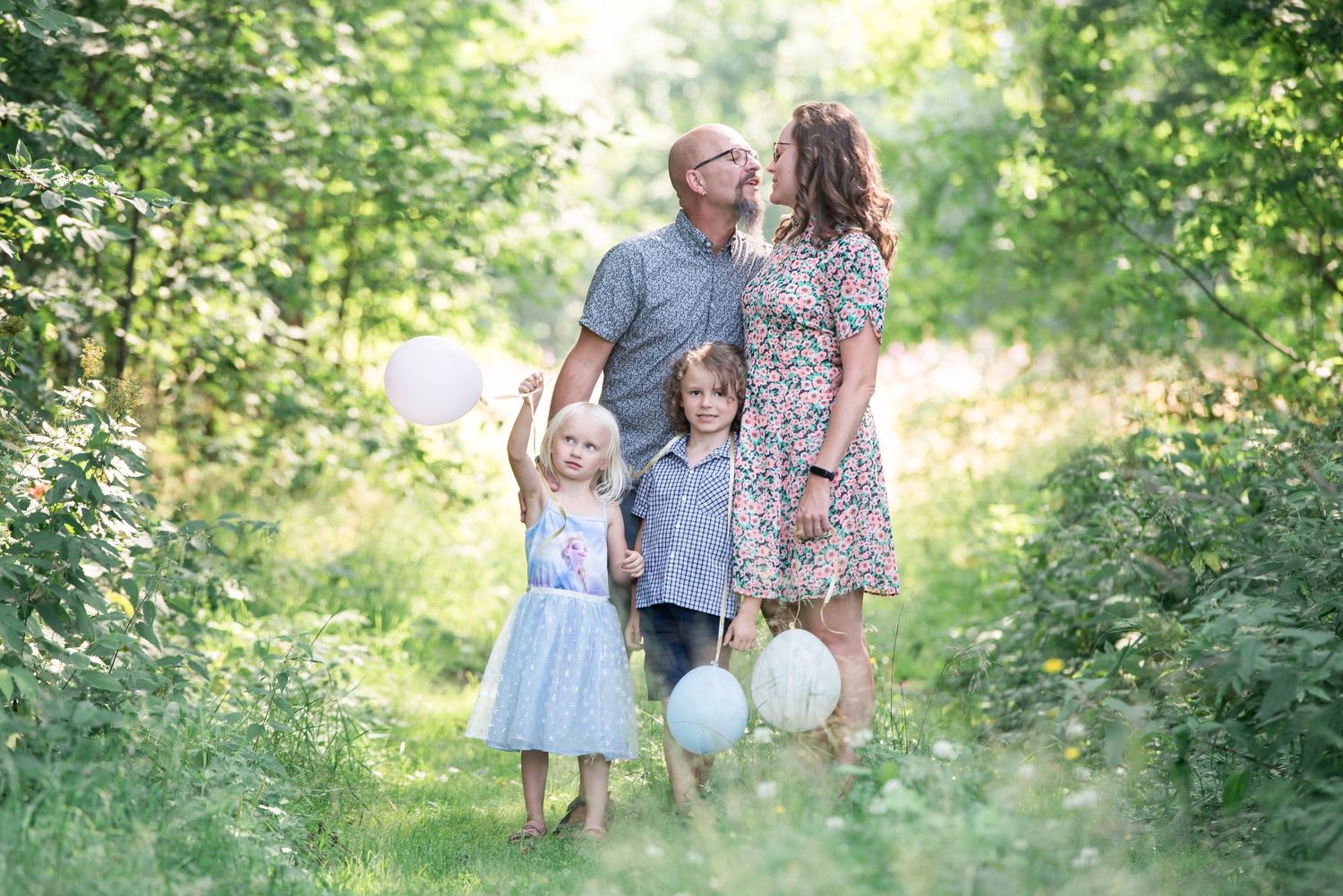 Family of four with balloons posing in a lush green forest path, parents sharing a kiss above two young children.