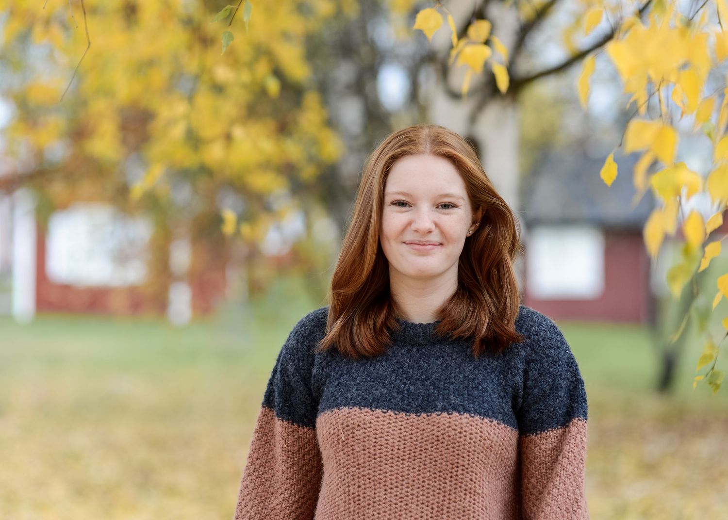 Young woman with red hair wearing a cozy sweater smiling outdoors among golden autumn birch trees.