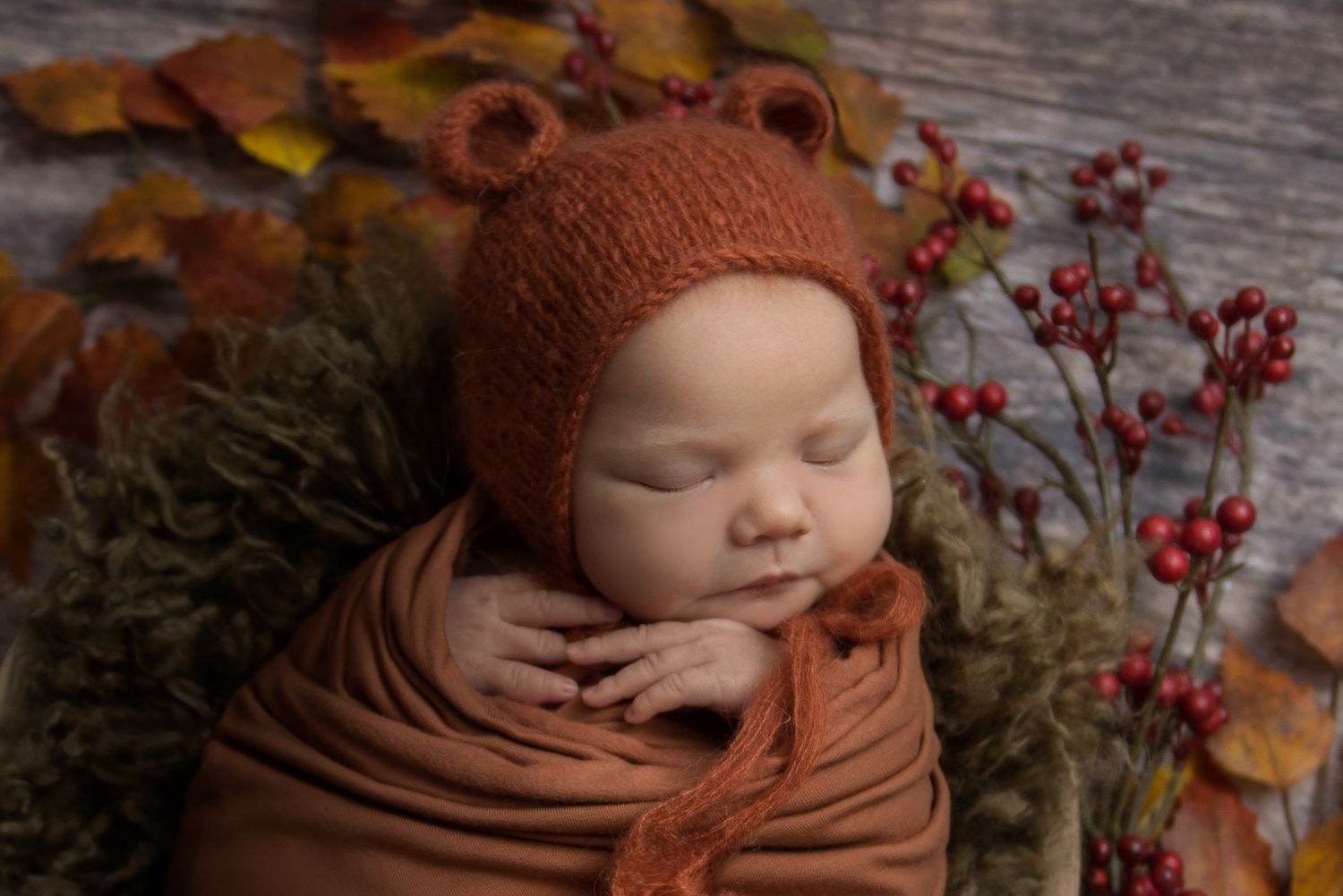 Autumn-themed newborn photo with baby wrapped in orange knits among fall berries and leaves.
