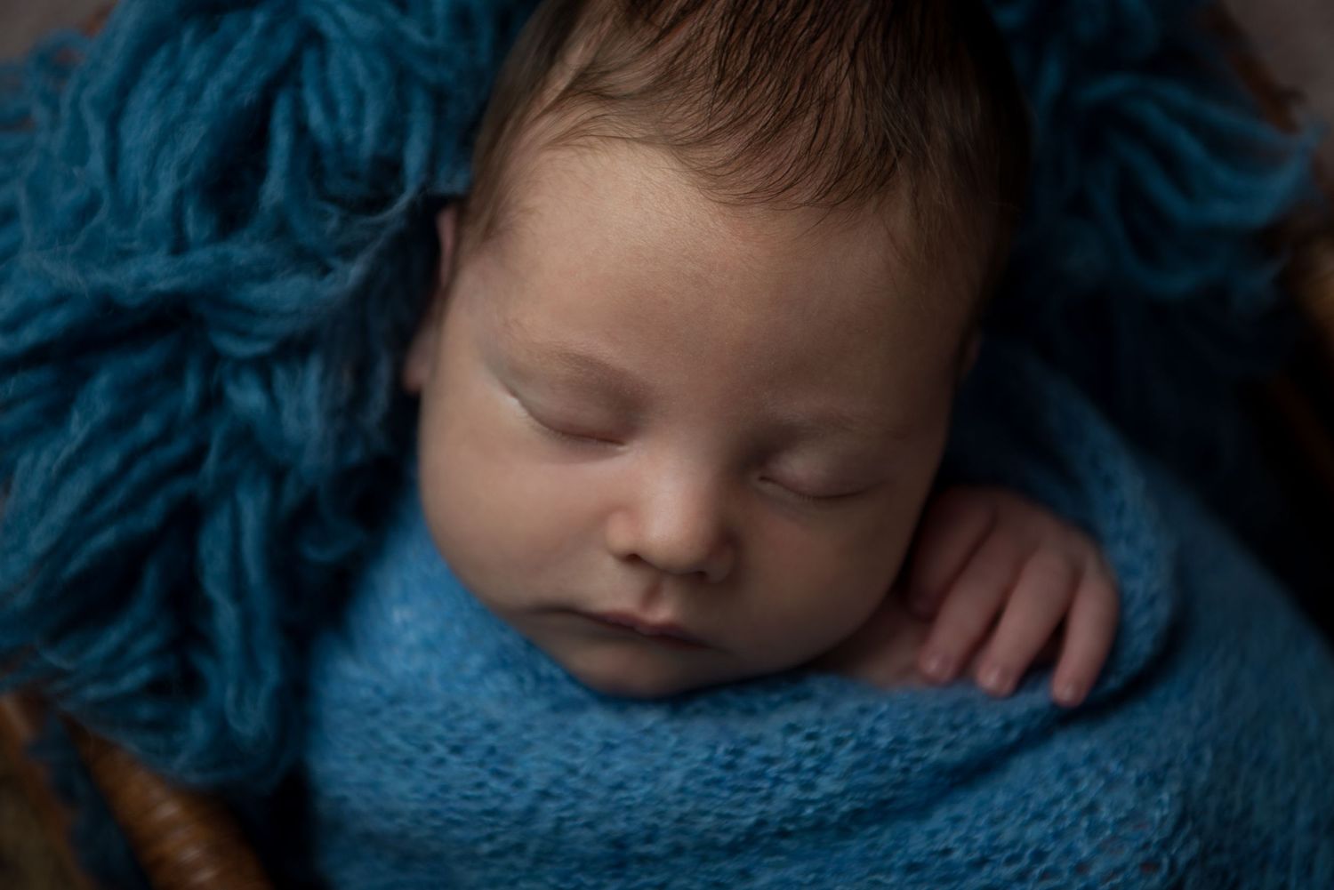 Sleeping newborn wrapped in blue knit blanket with matching background.