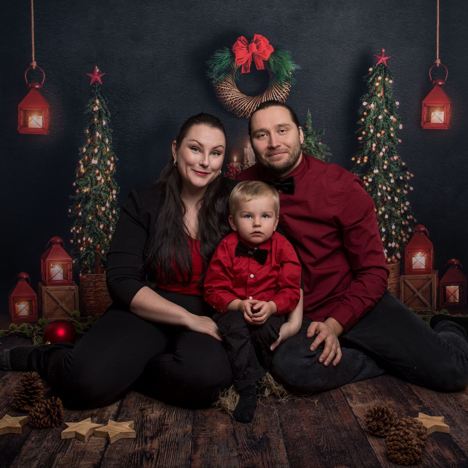 Festive holiday photo scene with Christmas trees, lanterns and pine cones on wooden floor.