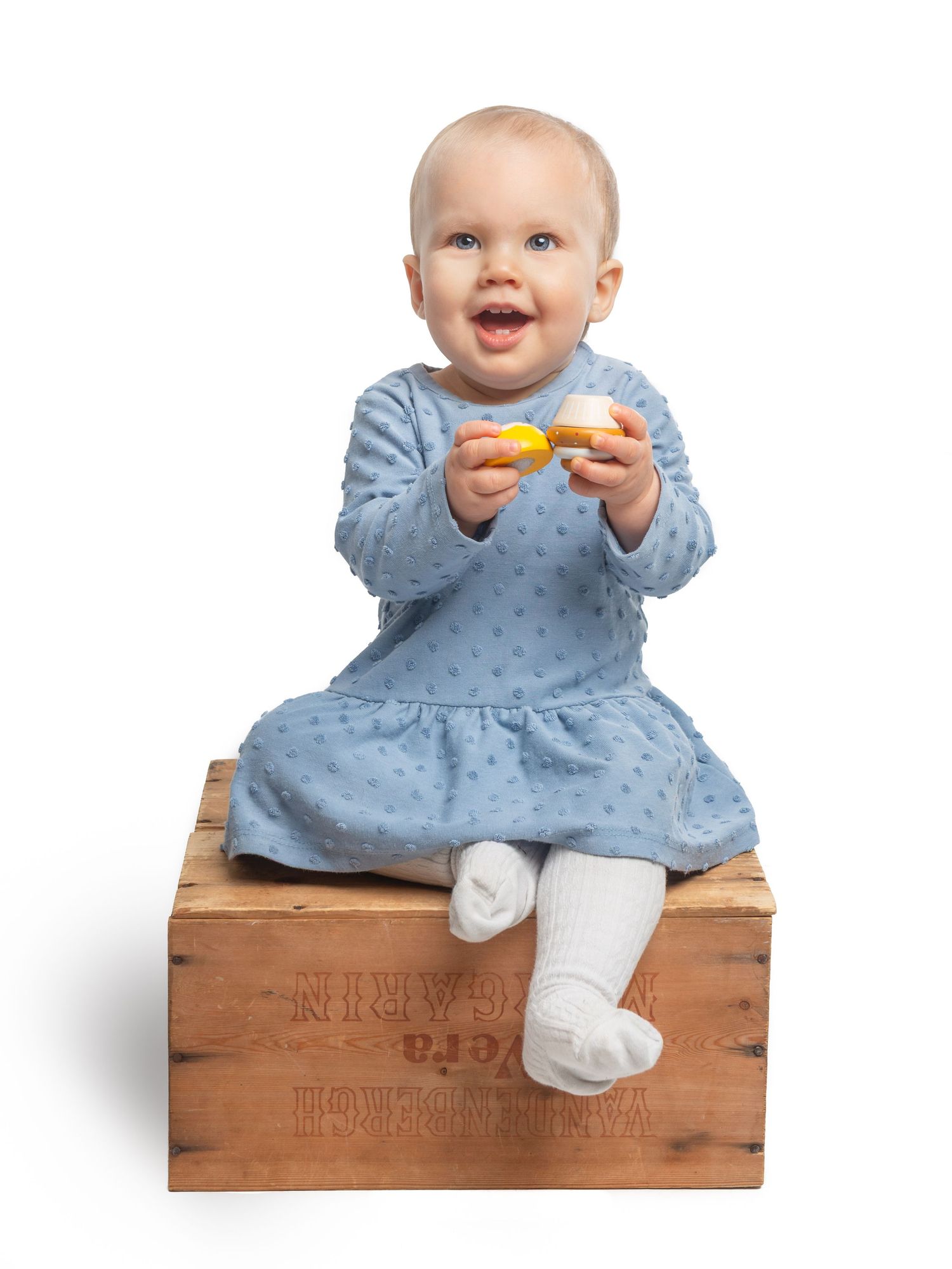 Baby in blue dress sitting on wooden crate against white background.