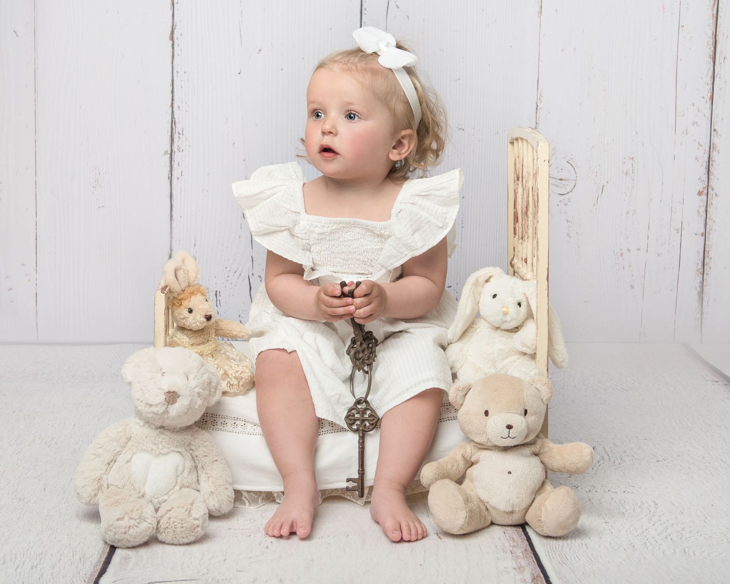 Child in white dress sits among plush teddy bears on white wooden floor.