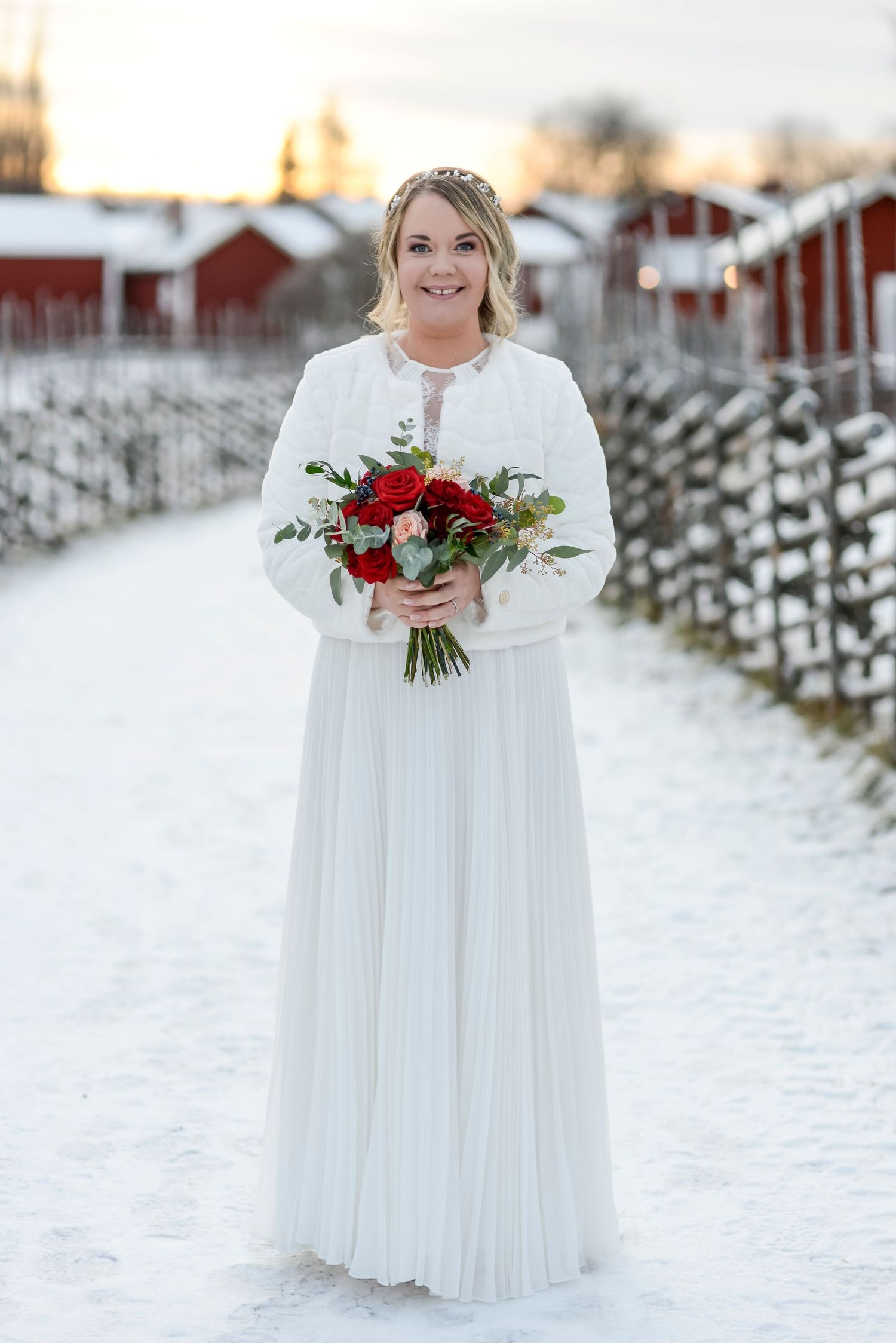 Winter bride in flowing white dress poses with red rose bouquet against snowy backdrop.