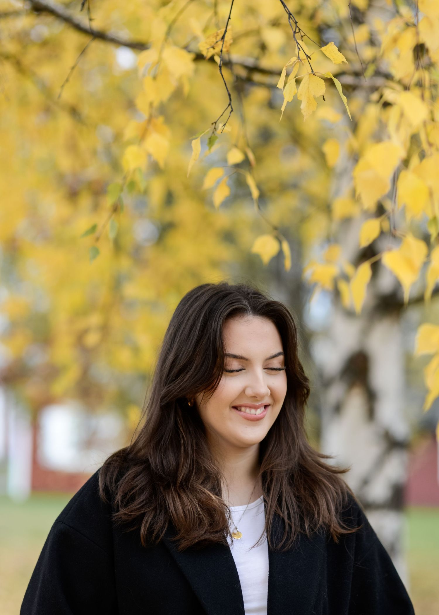 Young woman with dark hair smiling outdoors in autumn, wearing a black coat with yellow fall foliage in background.