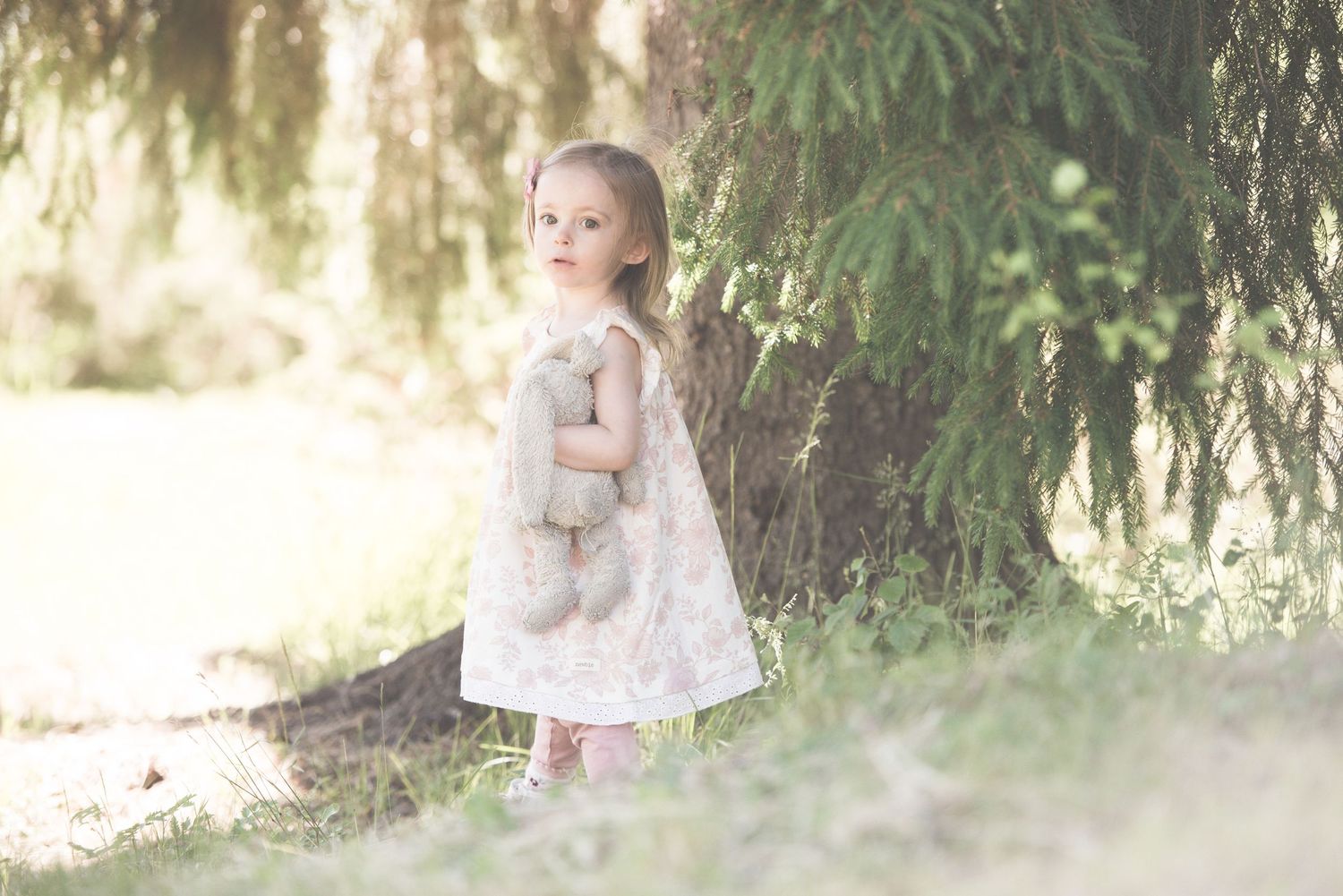 Young girl in floral dress holding teddy bear standing under weeping willow tree in soft natural light.