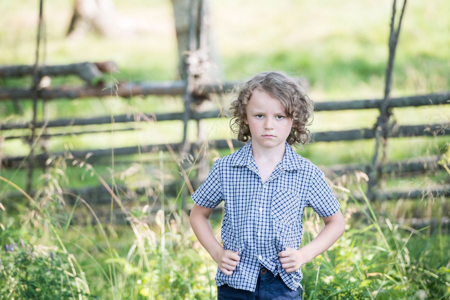 A young curly-haired boy in a blue plaid shirt stands confidently with hands on hips in a green rural field.