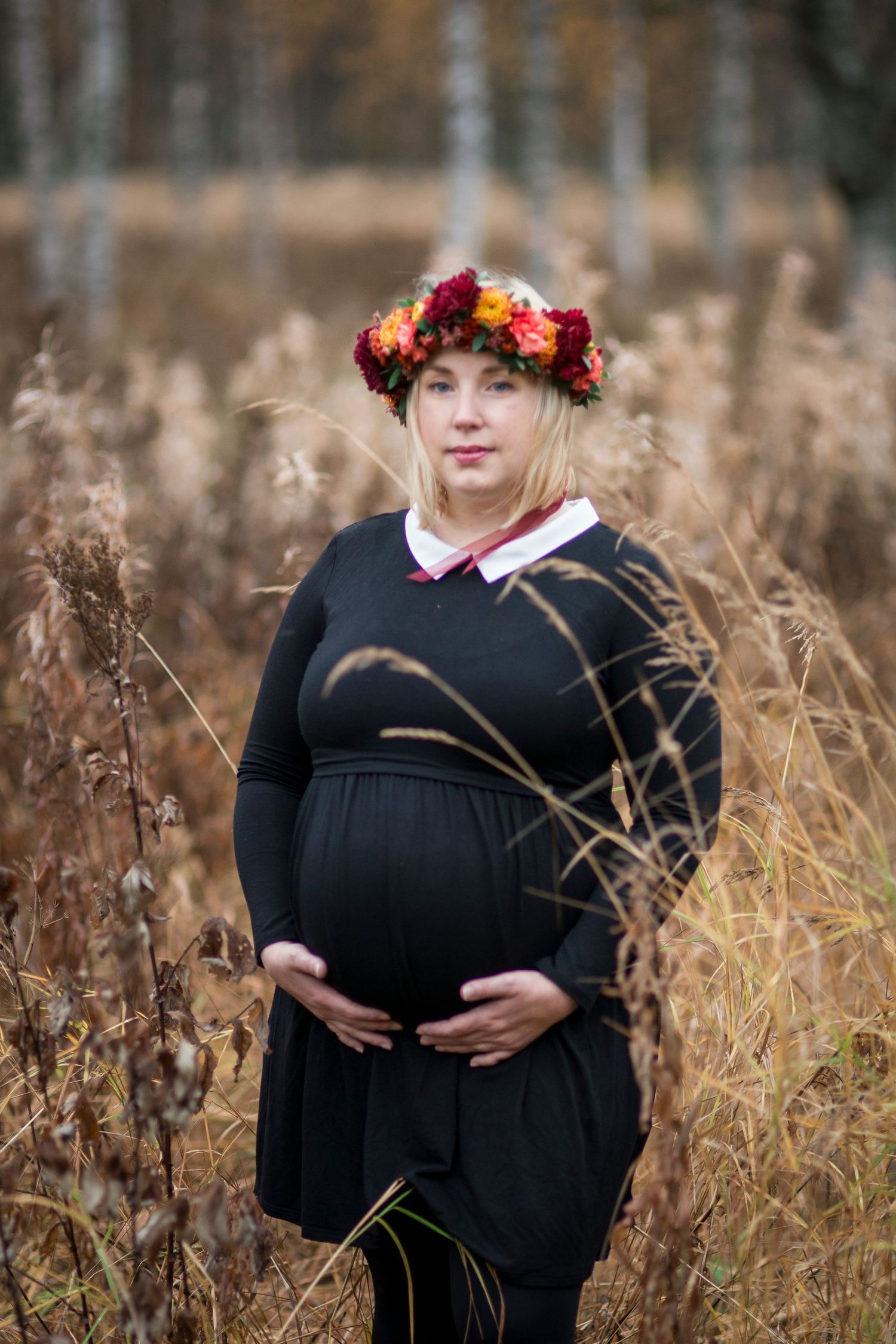 Pregnant blonde woman wearing floral crown and black dress standing in dry autumn grass field.