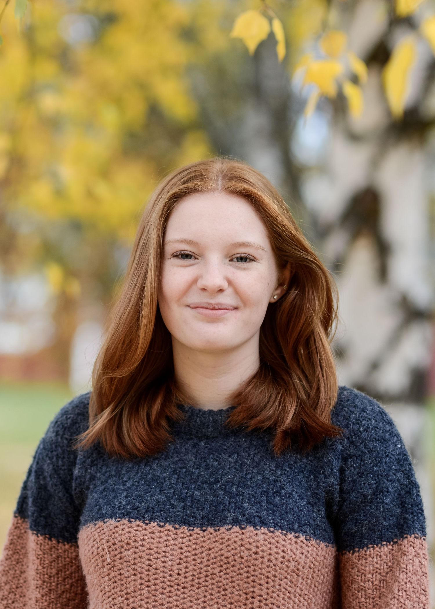 Young woman with red hair smiling outdoors in autumn, wearing a navy and pink striped knit sweater.