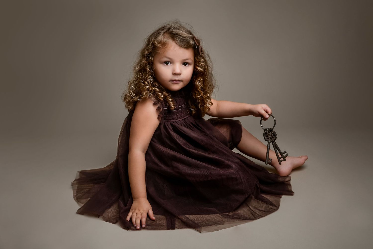 A young child in a brown dress poses gracefully in a studio against a neutral backdrop.
