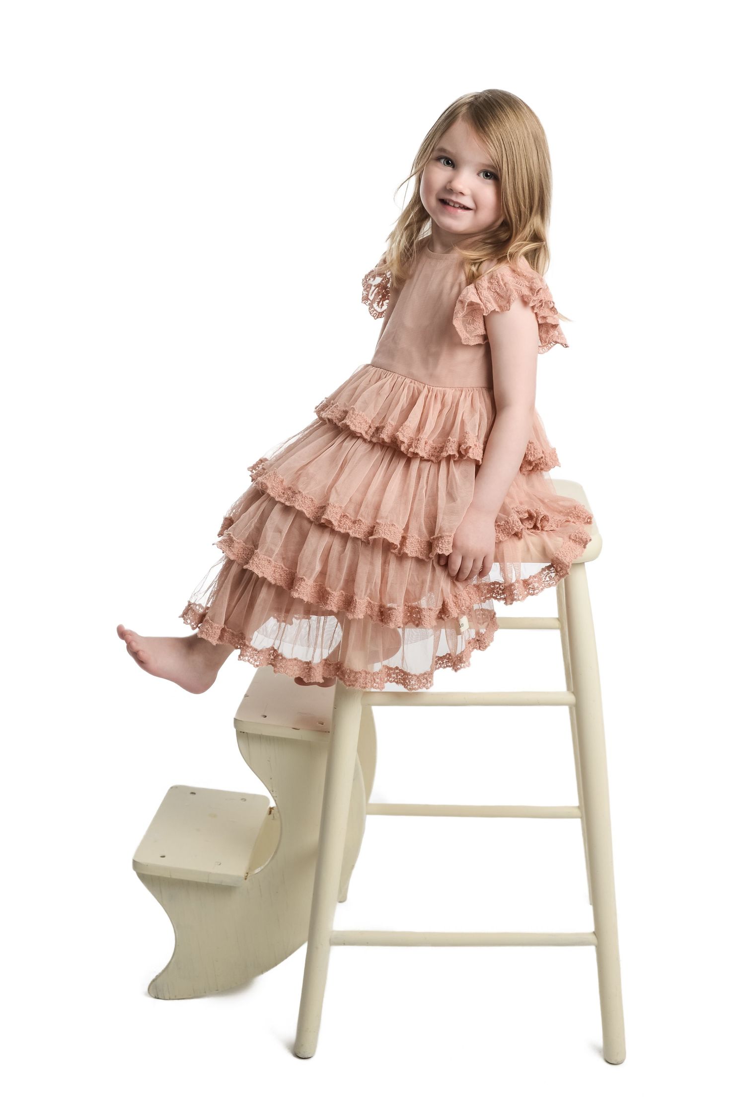 Child wearing peach ruffle dress sitting playfully on white chair.