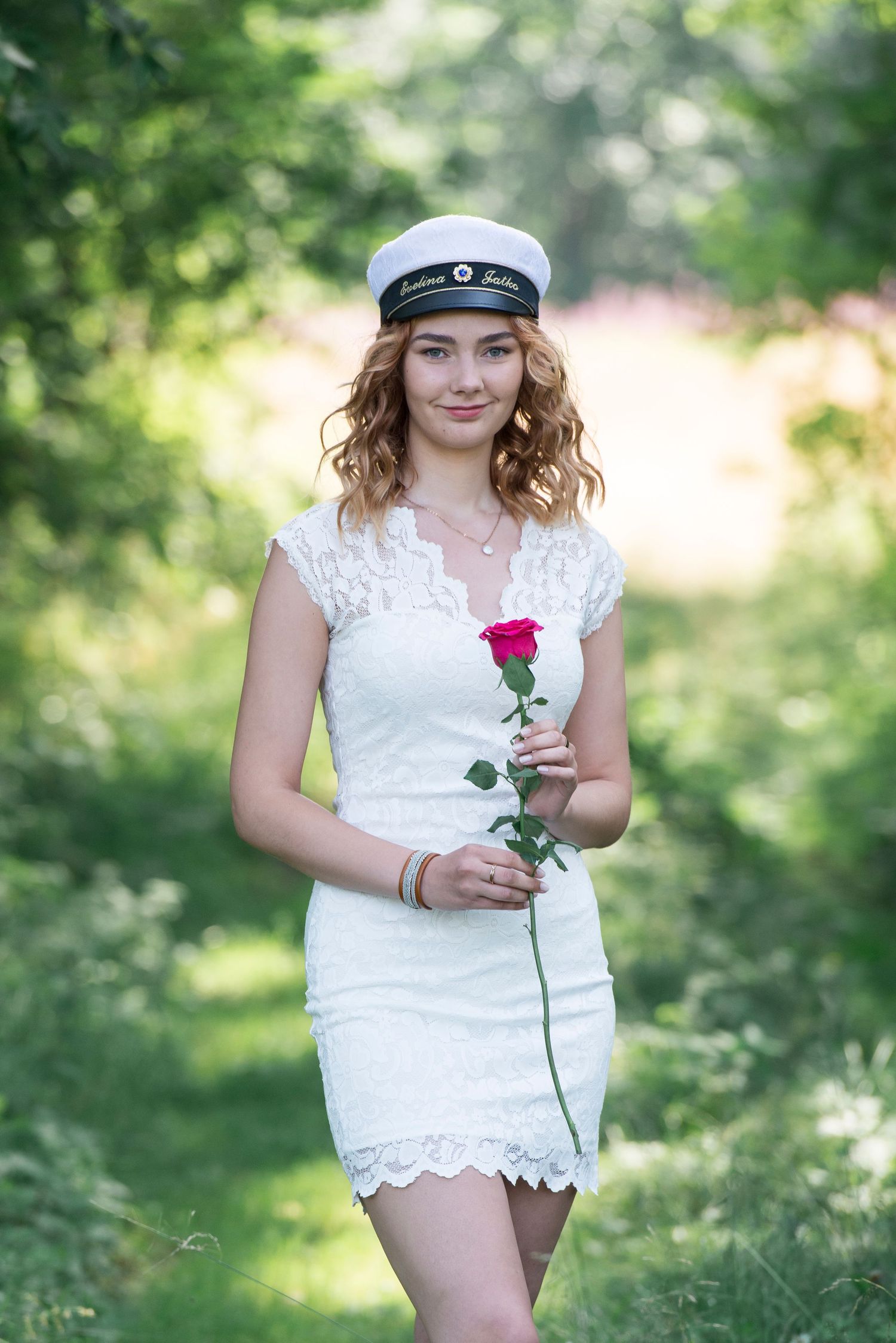 Figure in white lace holding a single red rose in a sunlit garden setting.