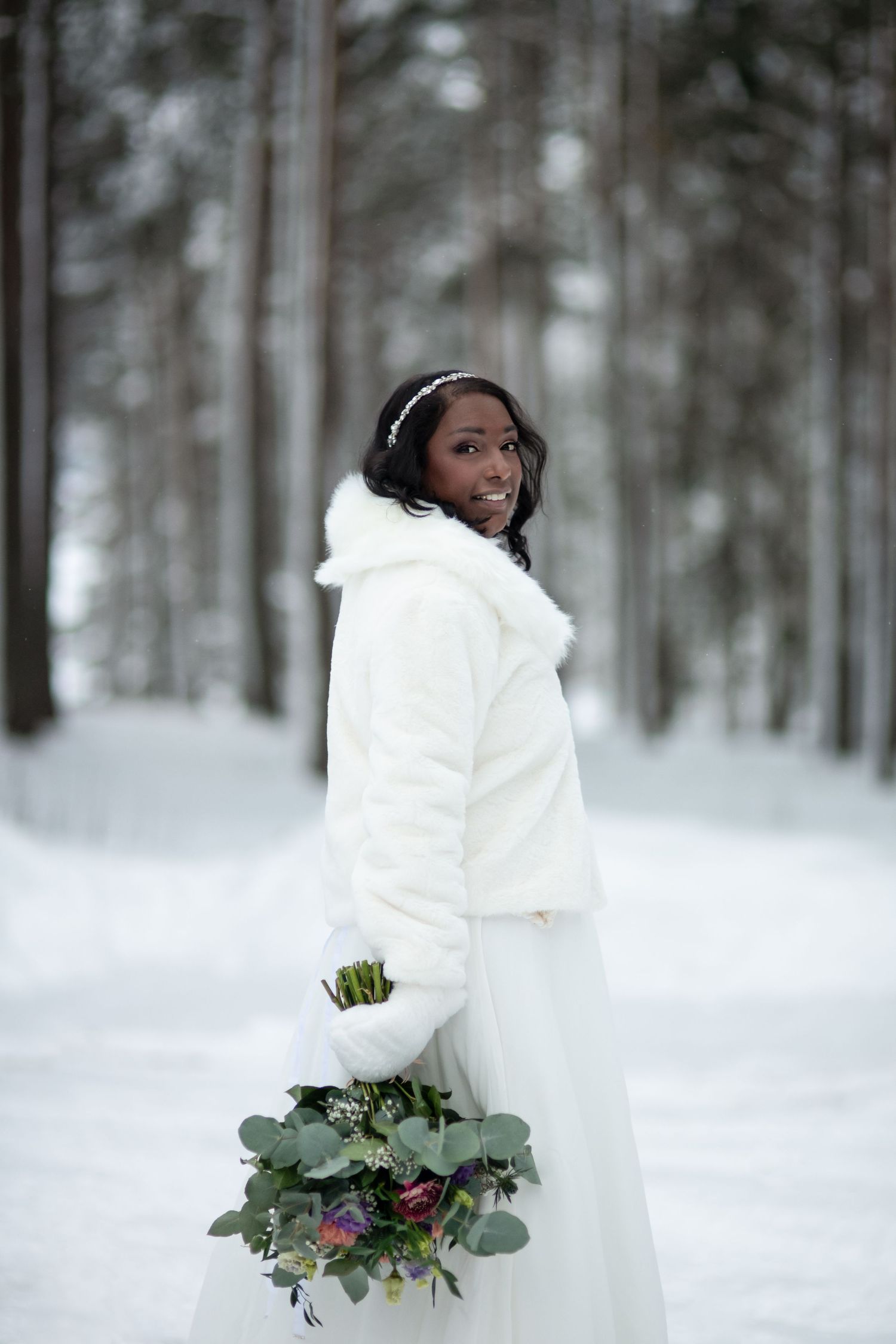 A bride in a white winter coat holds a bouquet while standing in a snowy forest setting.