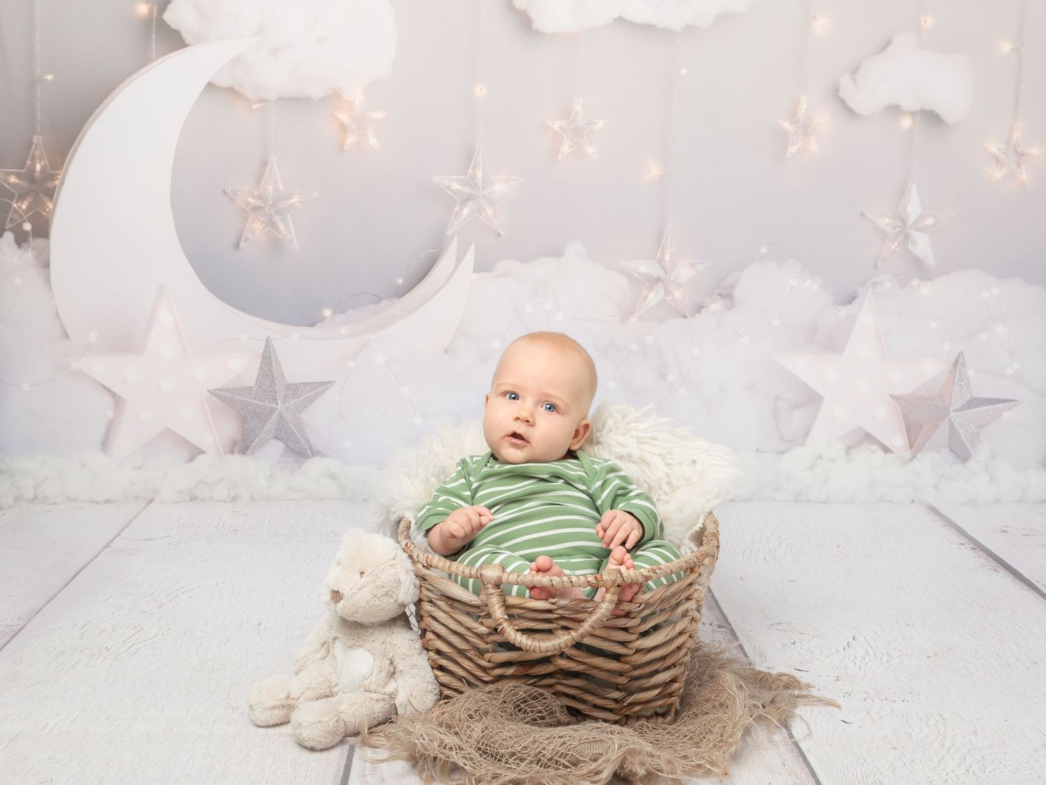 Baby in green striped outfit sits in wicker basket surrounded by stars and moon nursery backdrop.
