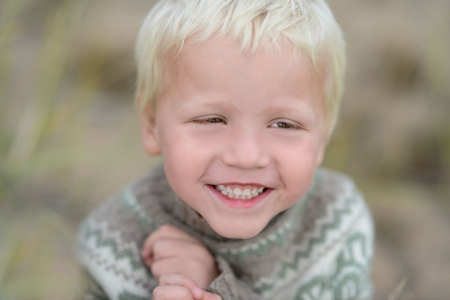 Smiling blond boy in a patterned sweater, hands clasped, with a joyful expression outdoors.