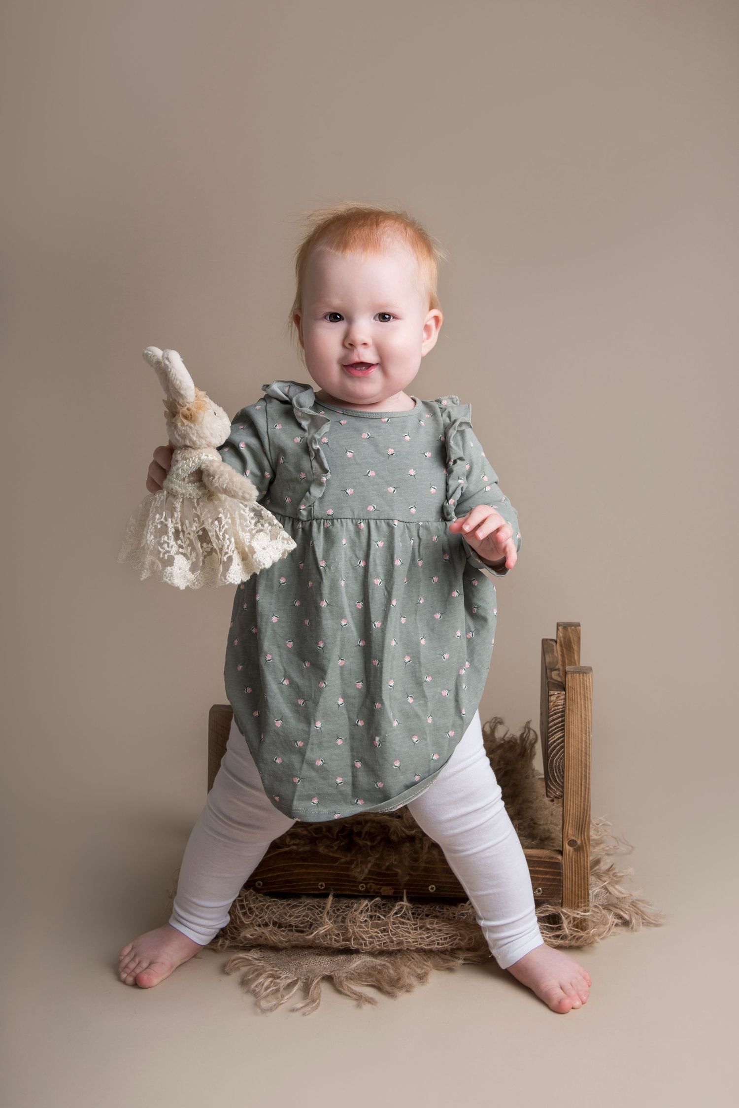 Baby in sage green dress sitting on wooden prop against neutral background.