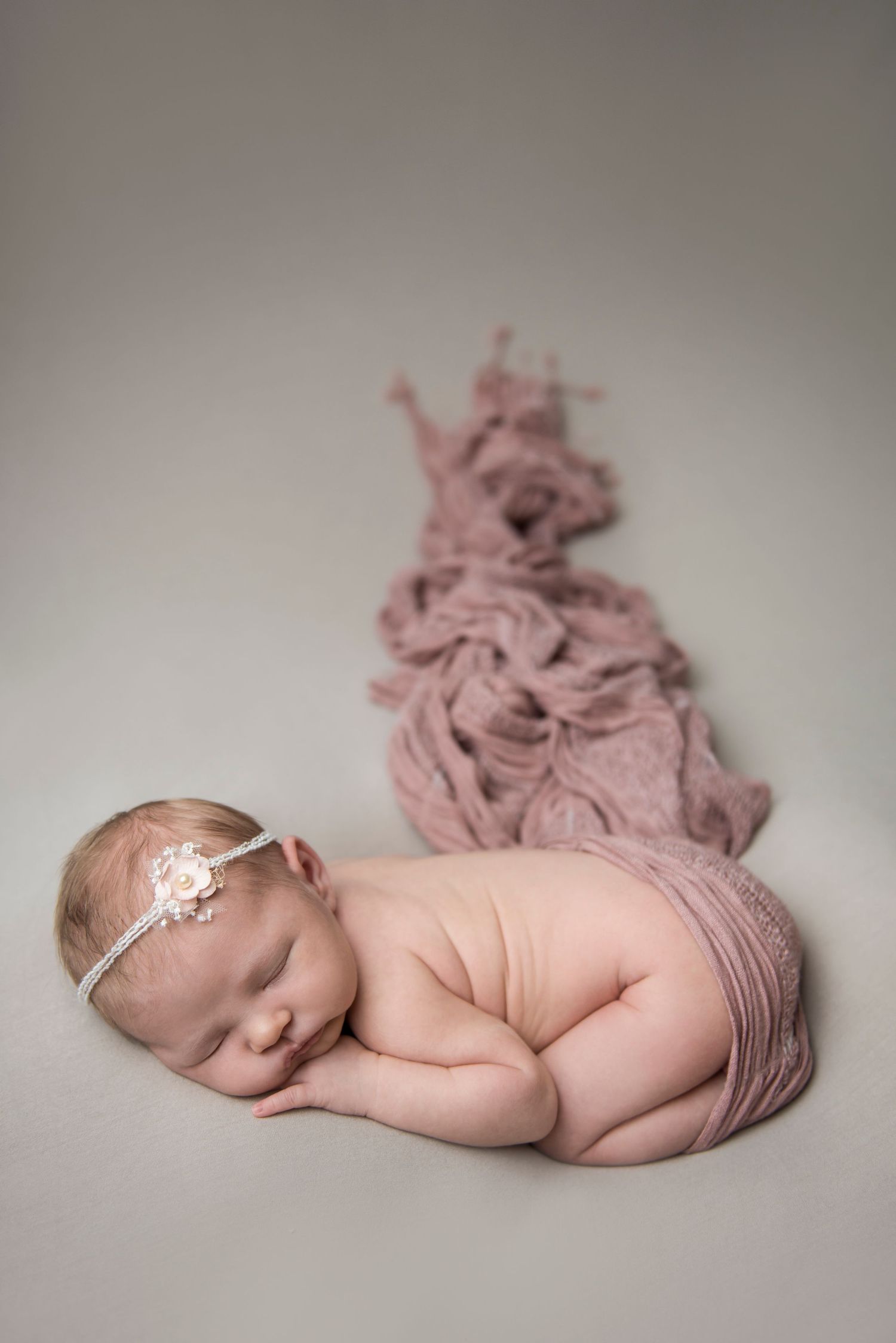 Sleeping baby with lace headband beside flowing pink fabric in studio setting.