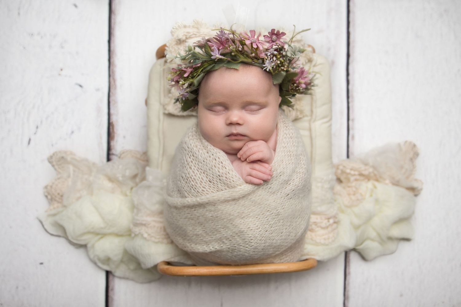 Newborn photography setup with cream colored wraps and delicate floral accents on white wooden backdrop.