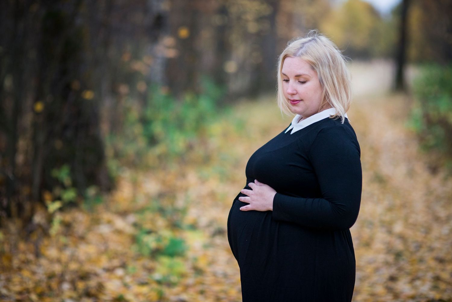 Pregnant woman in black dress cradling her baby bump in an autumn park with golden fallen leaves.