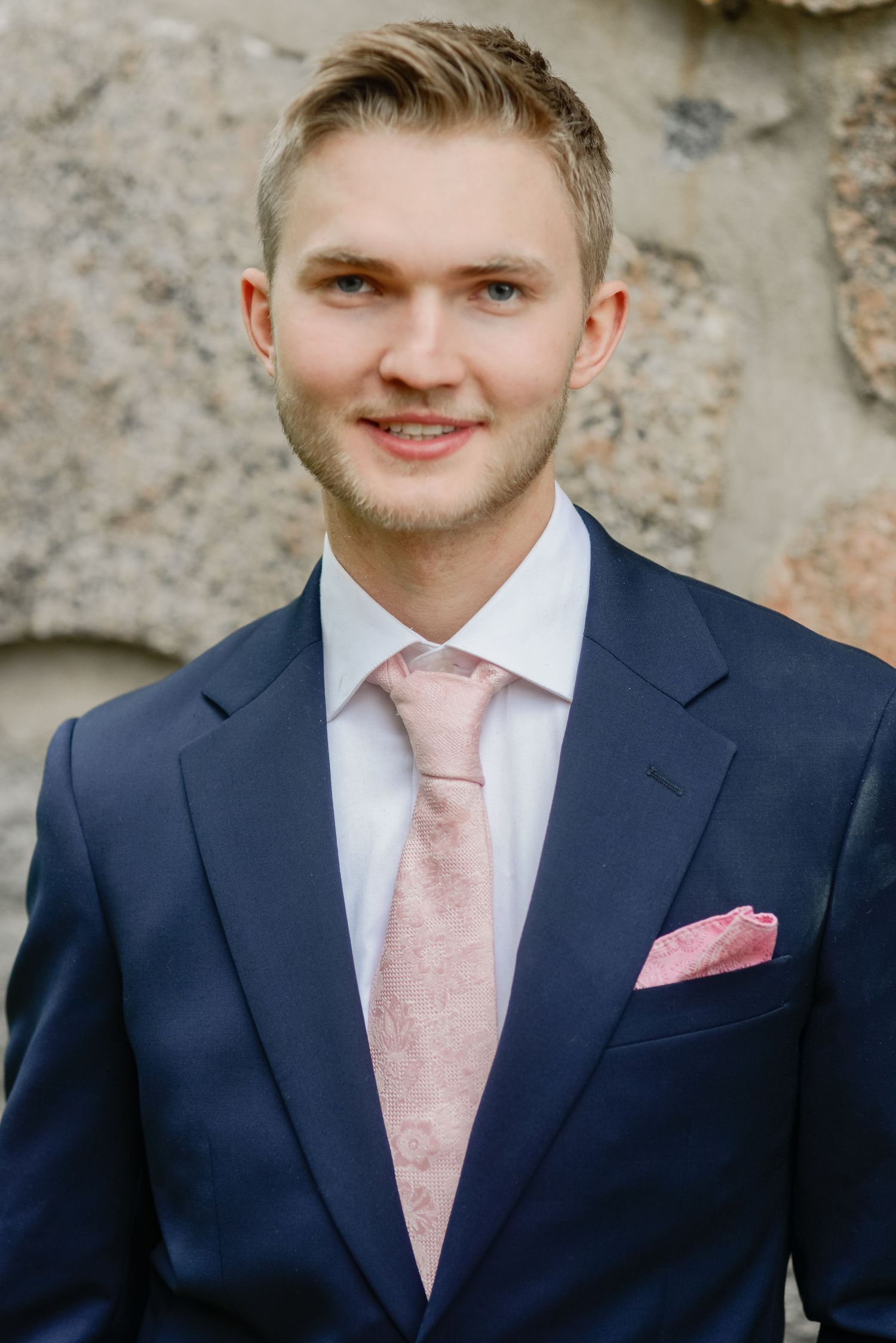 Portrait of person in navy suit with pink tie against textured background.