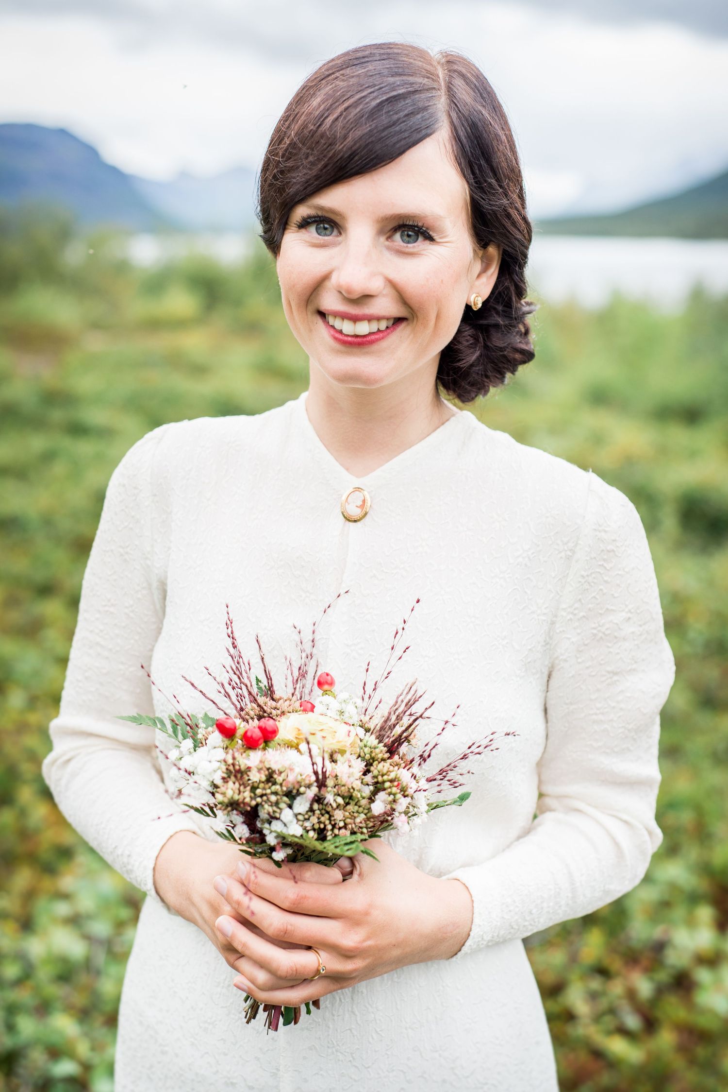 Bride in white dress holds delicate wildflower bouquet against mountain backdrop.