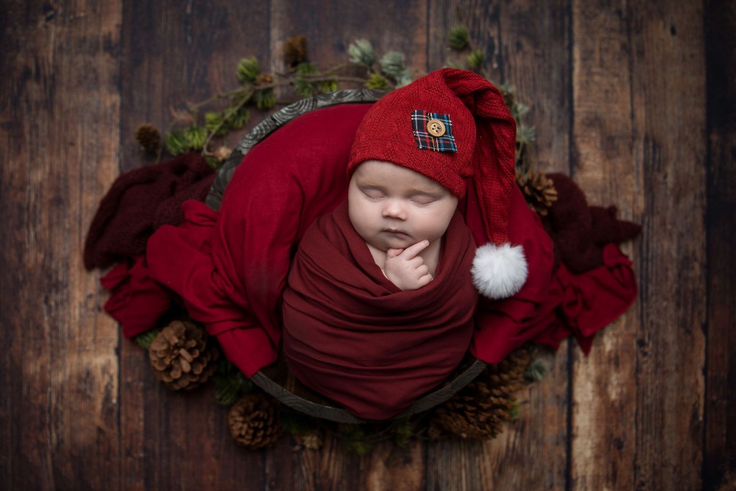 Holiday newborn photo with baby wrapped in deep red fabric on rustic wooden background.
