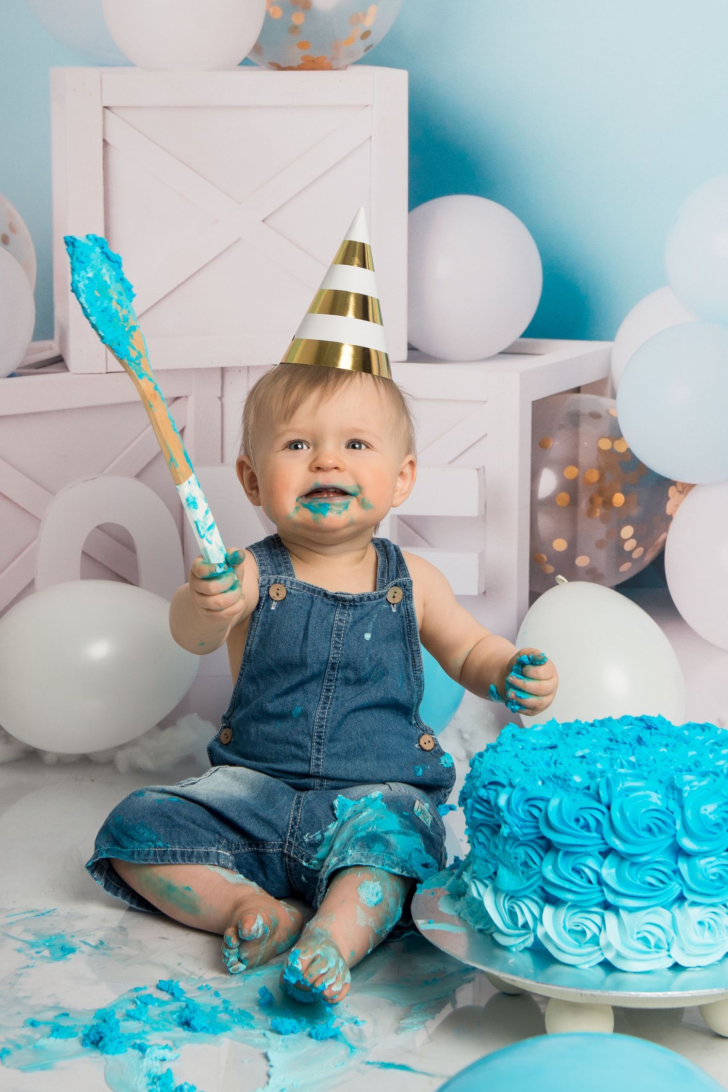 A first birthday cake smash scene with blue frosting, gold party hat, and festive balloon decorations.