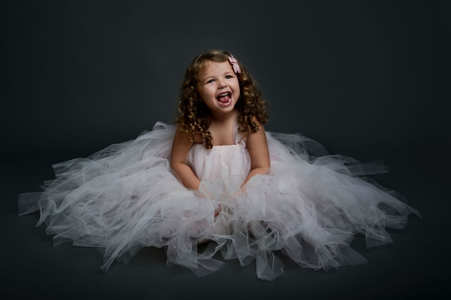 Young girl in white tulle dress laughing joyfully against dark background in professional studio portrait.