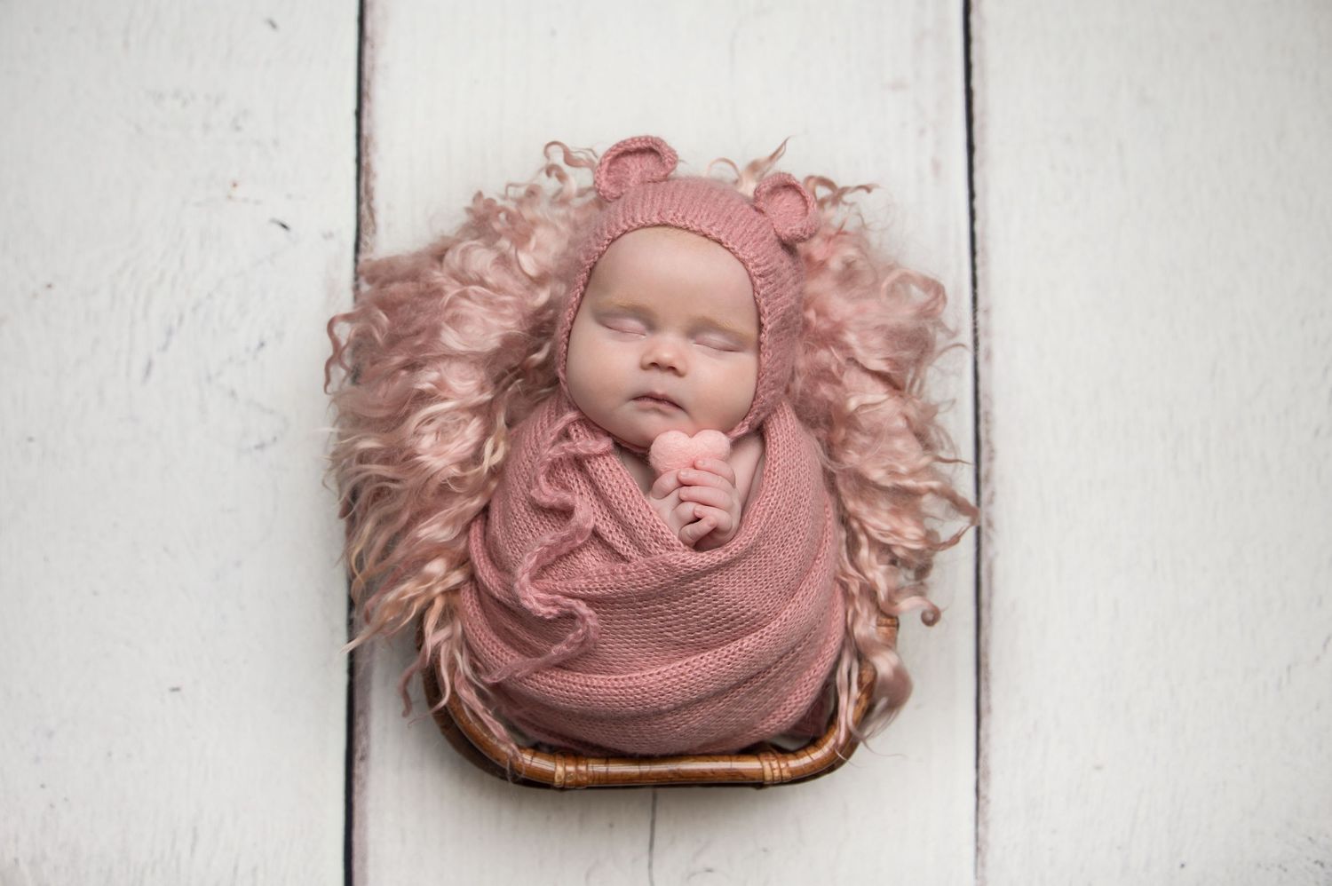 Newborn photography with baby wrapped in soft pink fabric on white wooden background.