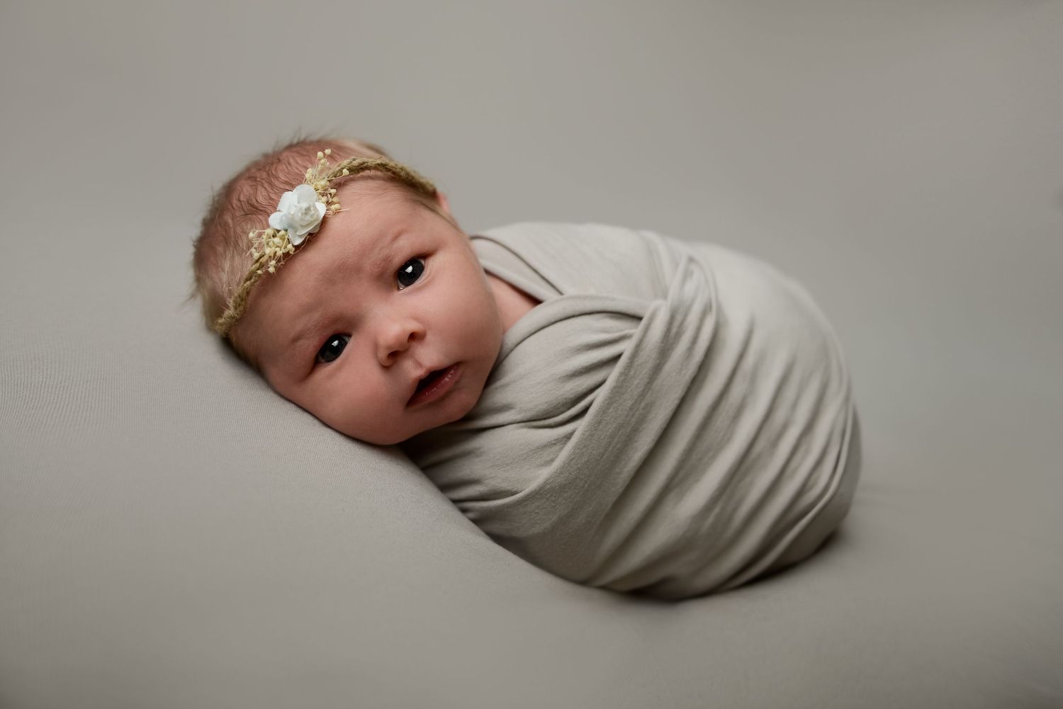 Newborn wearing delicate headband wrapped in light fabric against grey backdrop.