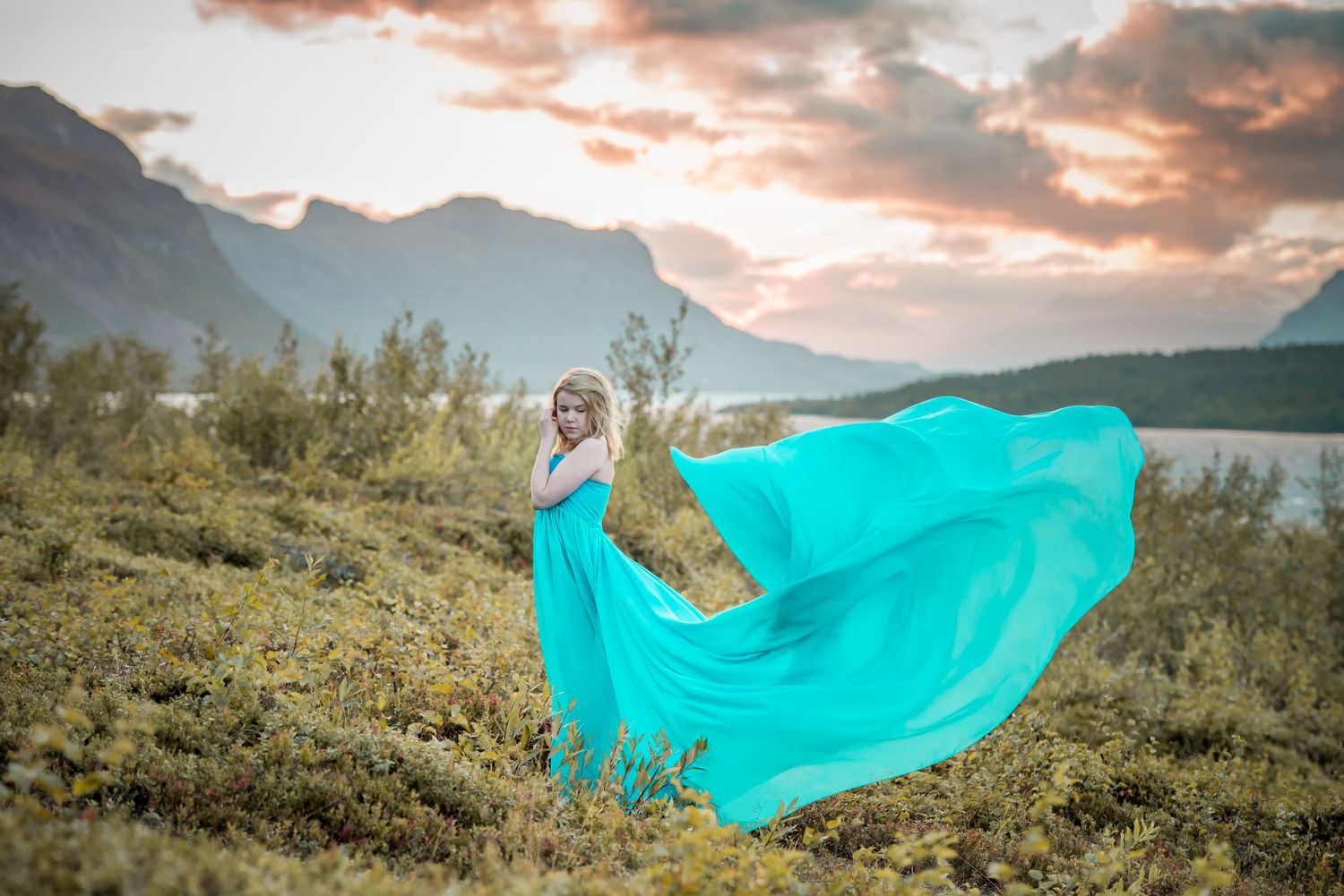 Blonde woman in billowing teal dress poses on hillside with mountain lake and sunset clouds behind her.